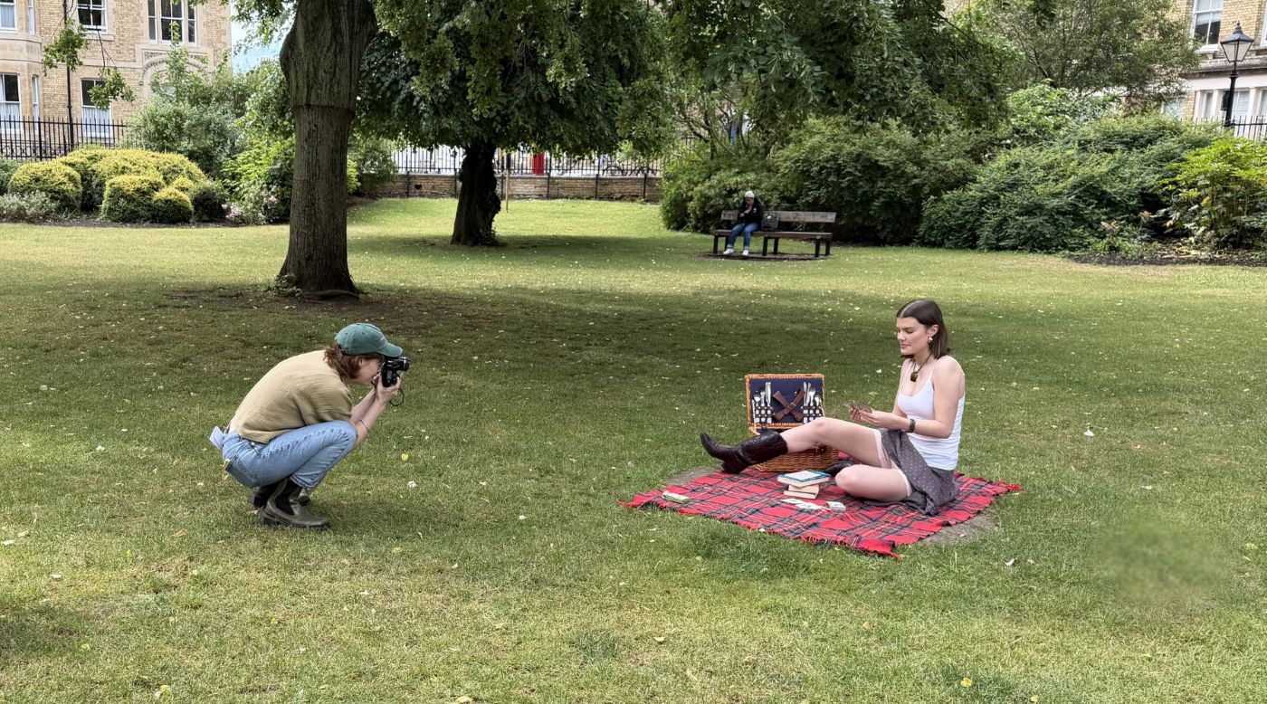 Wide angle photo of a park. In the centre to the left a photographer crouches to take a photo of a model (centre right) who sits on a red picnic blanket, wearing a skirt and boots.