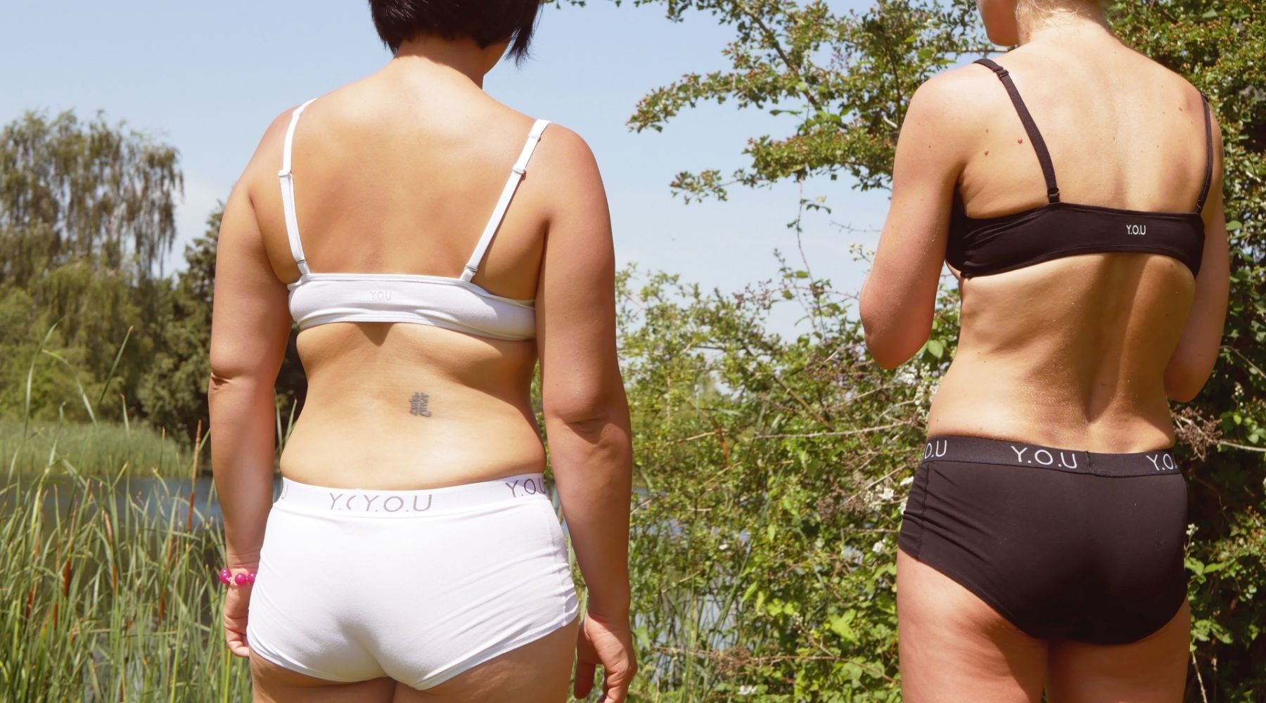 Photo of two women visible from behind as they stand looking out over a lake with greenery all around. One woman wears the Y.O.U Underwear bralette and Y.O.U waistband boy shorts in white while the other woman wears the black set.
