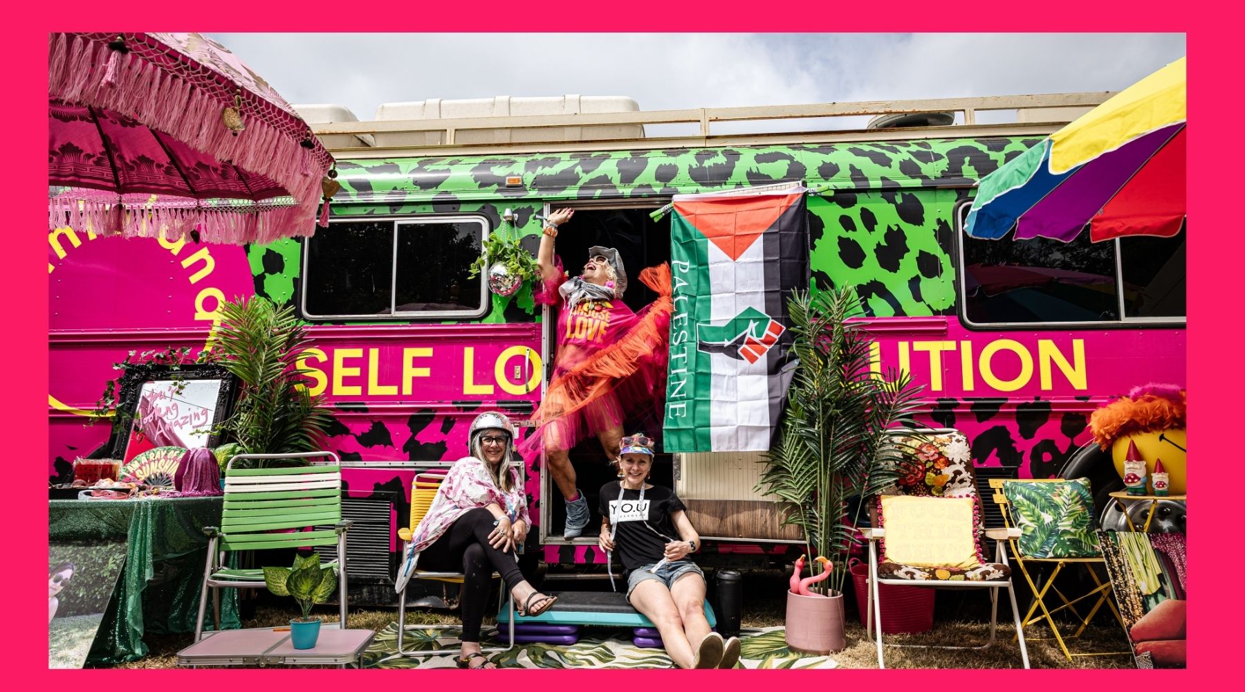 photo of three women posing outside a brightly decorated bus surrounded by chairs, plants, and parasols.