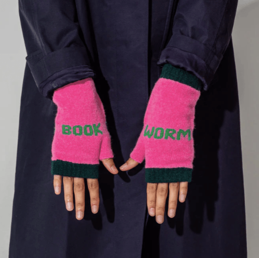 A close up photo of a model wearing the Black & Beech Book Worm mittens in bubble gum pink with green writing. The model is wearing a dark purple trench coat and standing in front of a beige background.