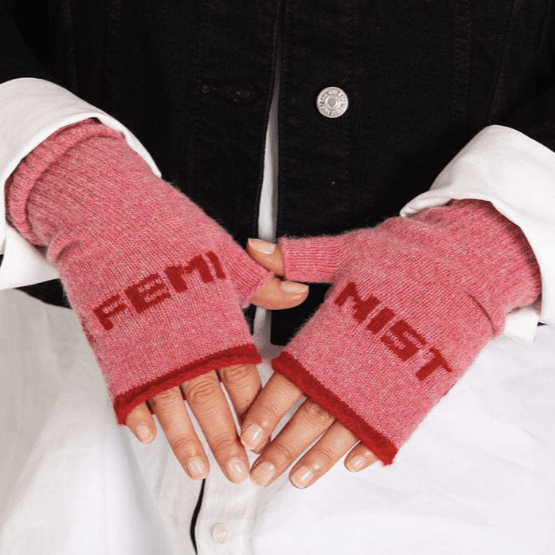 A close up photo of a model wearing the Black & Beech Feminist gloves in bubblegum pink with red writing and cuffs. The model is also wearing a long, over-sized white shirt with a black jacket.