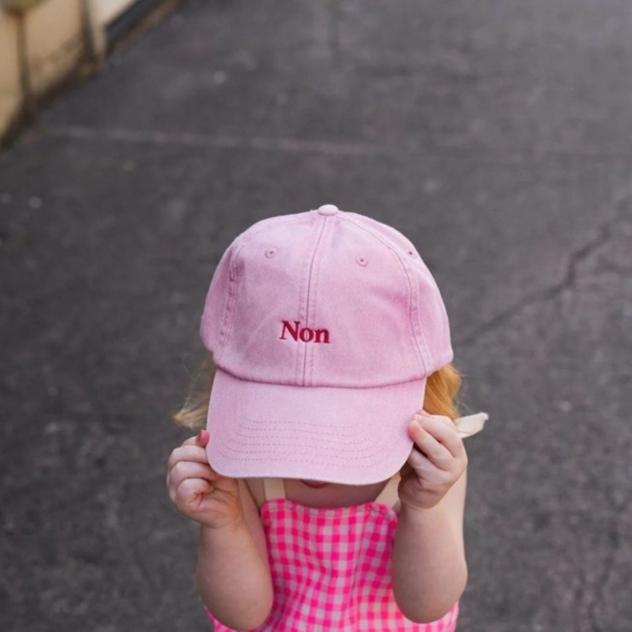 A photo of a young girl wearing the Black & Beech 'Non' baseball cap in pink, it is very oversized and she is standing on a concrete road