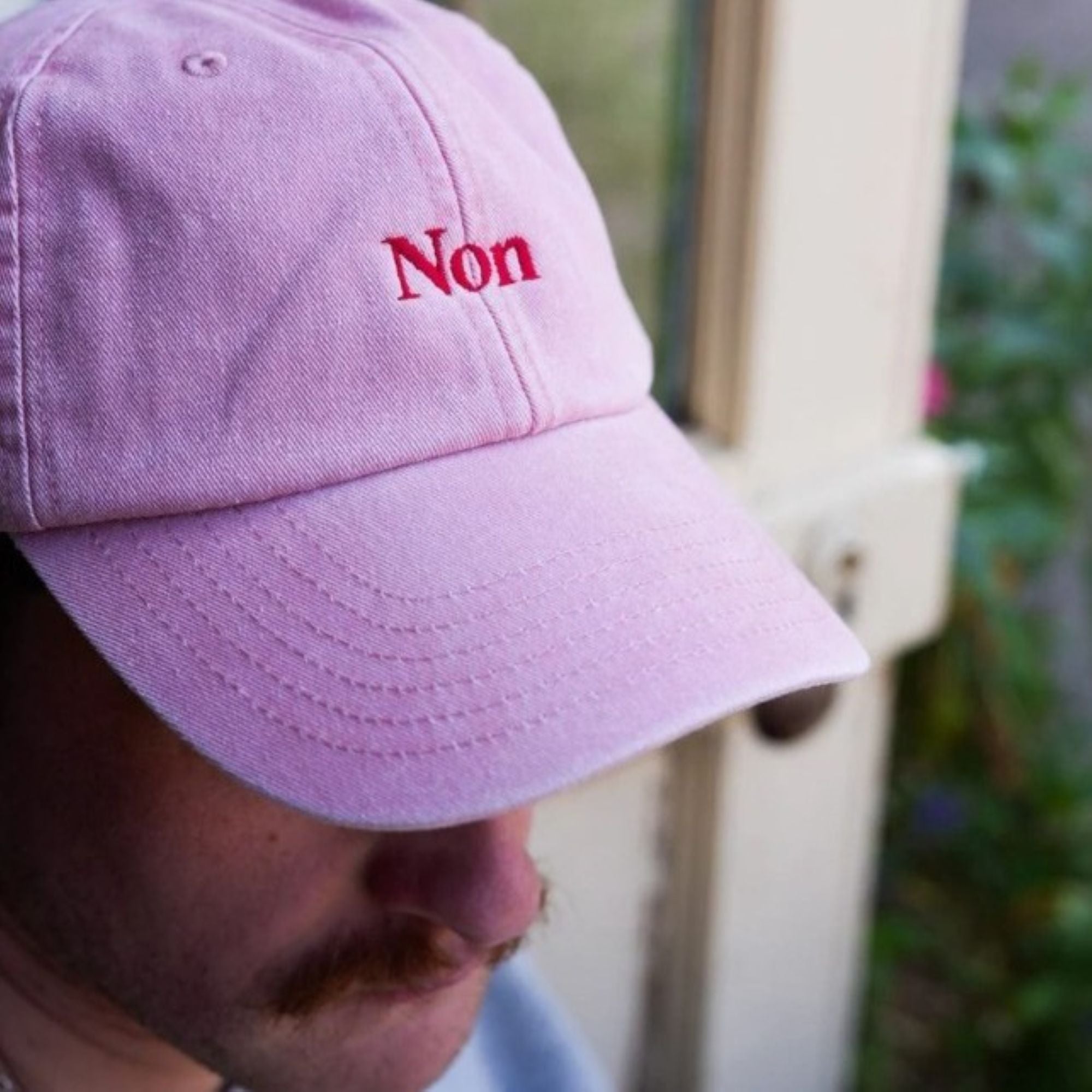 A photo of a man wearing the Black & Beech 'Non' baseball cap in pink standing near a garden door