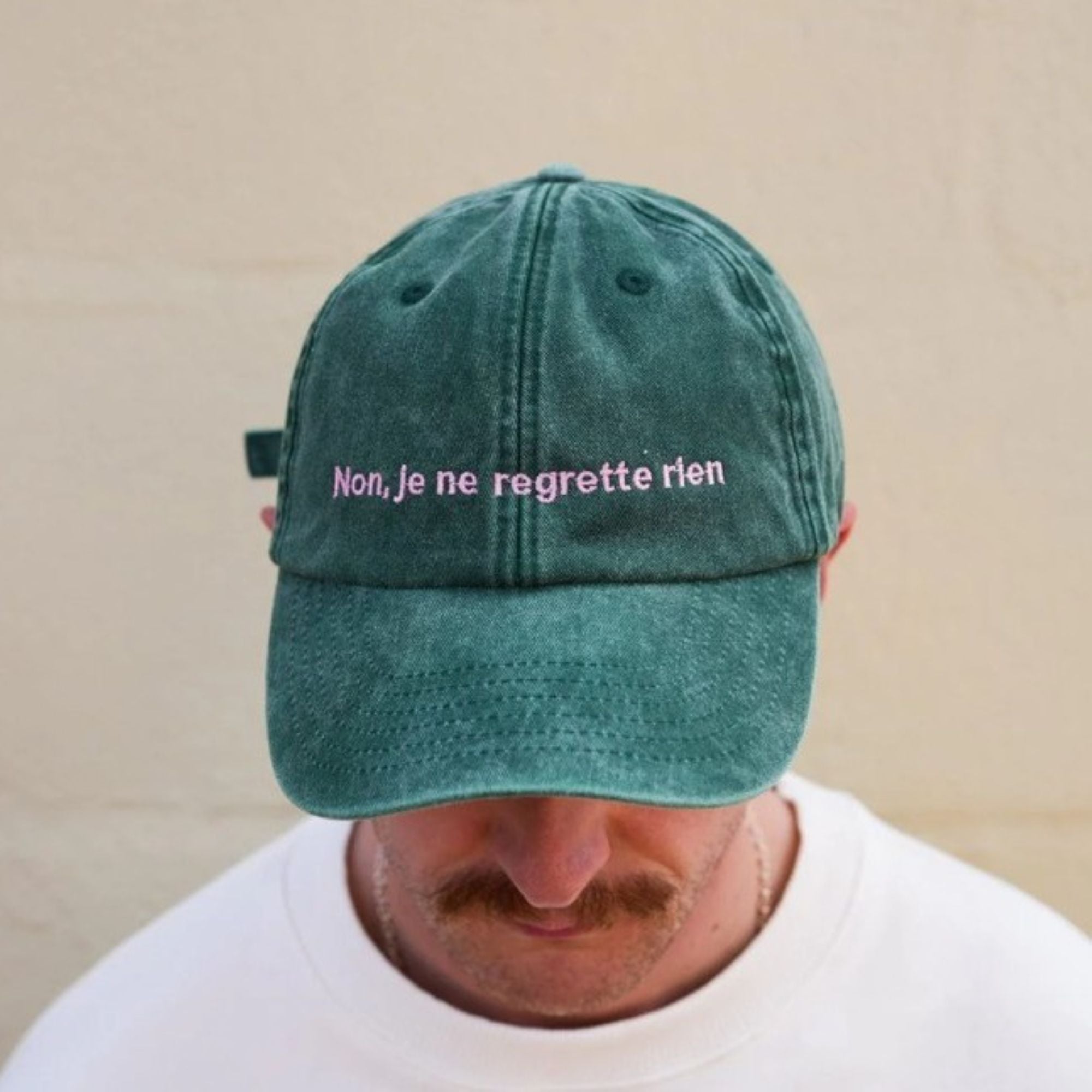 A mac wearing the Black & Beech Non Regrette baseball cap in forest green with baby pink writing. He is looking down so you can't see his eyes and standing in front of a cream background