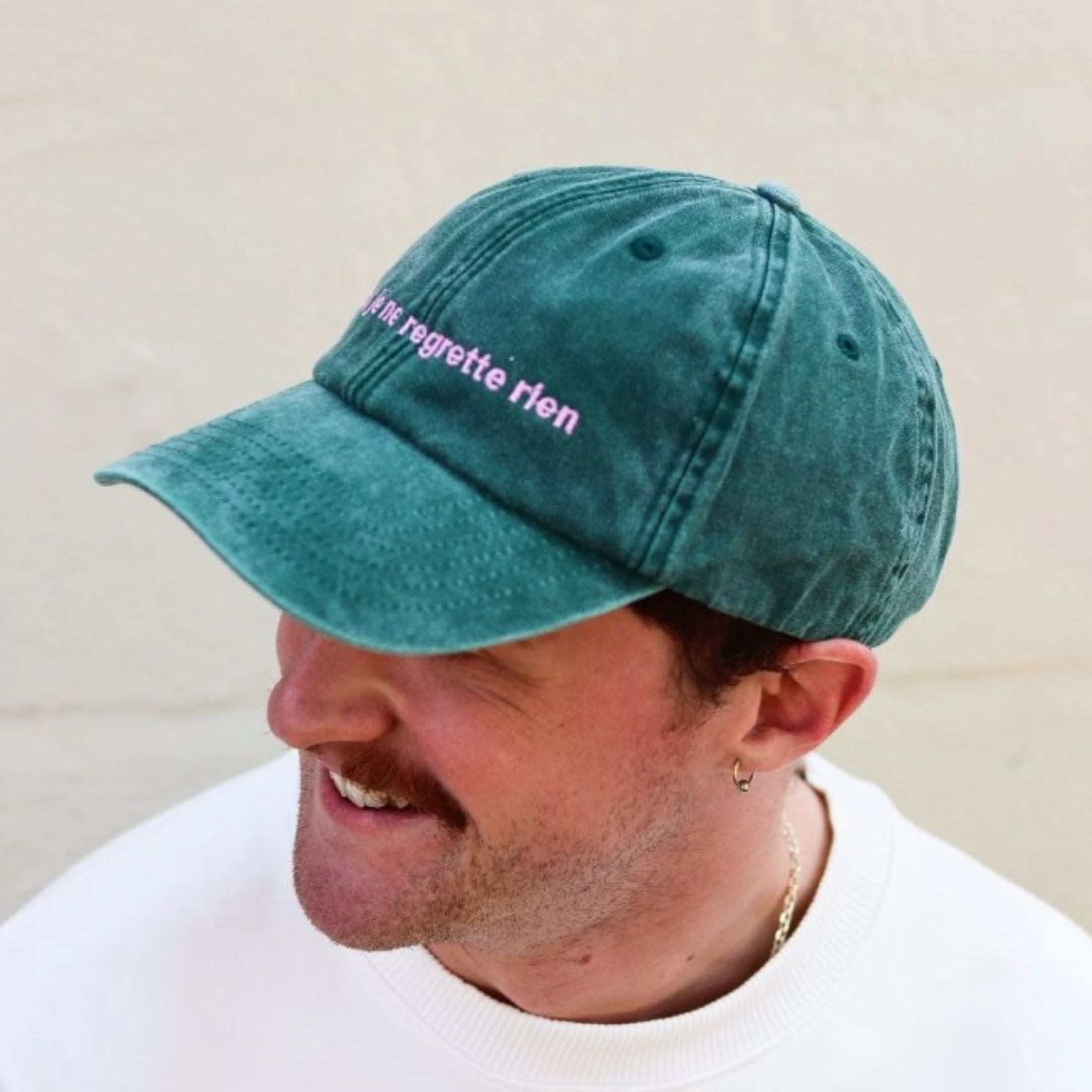 A man wearing the Black & Beech Regrette Cap in green, smiling and standing in front of a cream background