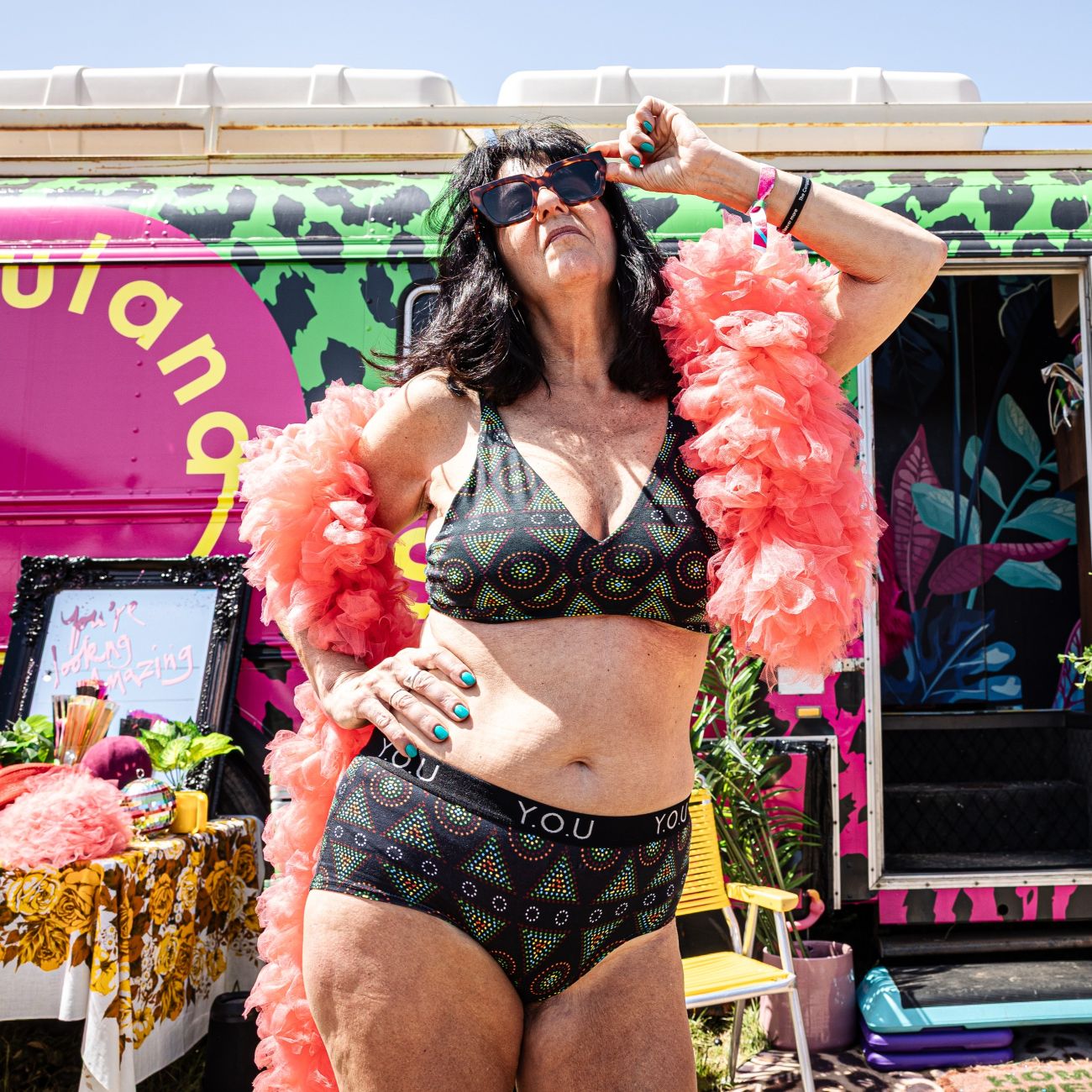 A photo of a model wearing the YOU Underwear bralette and branded boyshorts in the black mara print. She is standing in front of a bright green and pink coloured bus, facing the camera. She has a pink feather bower draped across her arms and big brown sunglasses. She is standing with her right hand on her hip and the other adjusting her sunglasses.