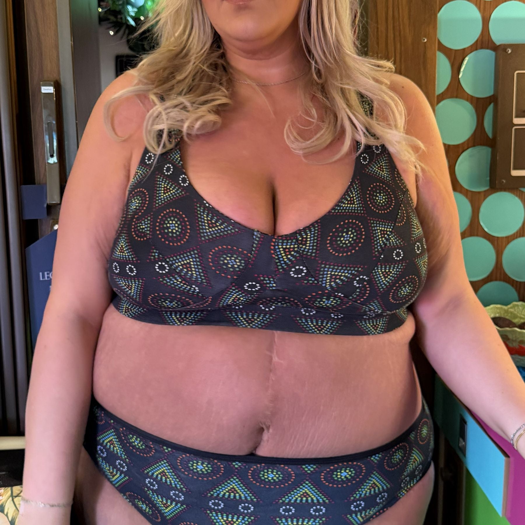 A photo of a model wearing the YOU Underwear black mara non-wired bra and matching knickers. The photo is zoomed in so we can see from the model's shoulder's to their hips. Behind them is a wooden wall with bright blue polka dots on. They are standing, facing the camera with their hands at their sides.