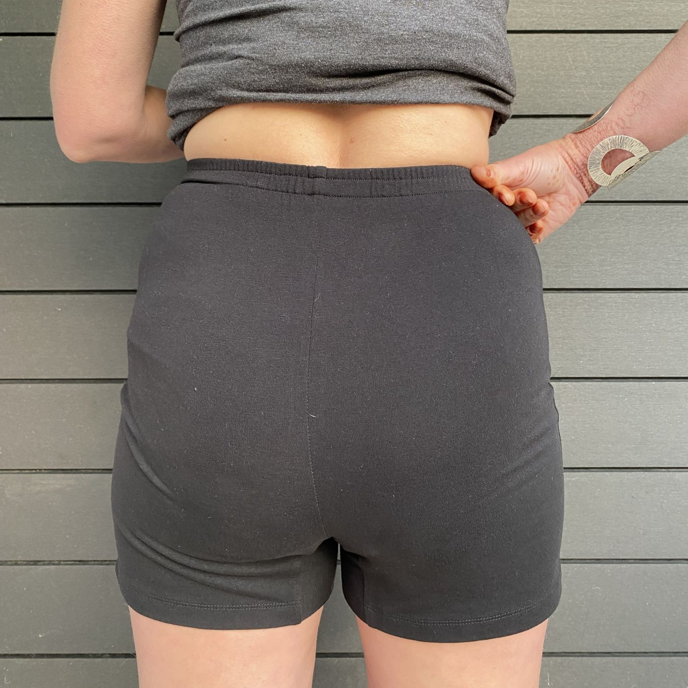 A close up photo of a model wearing the YOU Underwear undershorts in black. She is facing away from the camera, her right hand touching the waist of the short. There is a grey paneled background and the photo is zoomed in so we can see from the model's mid-stomach to their mid-thigh.