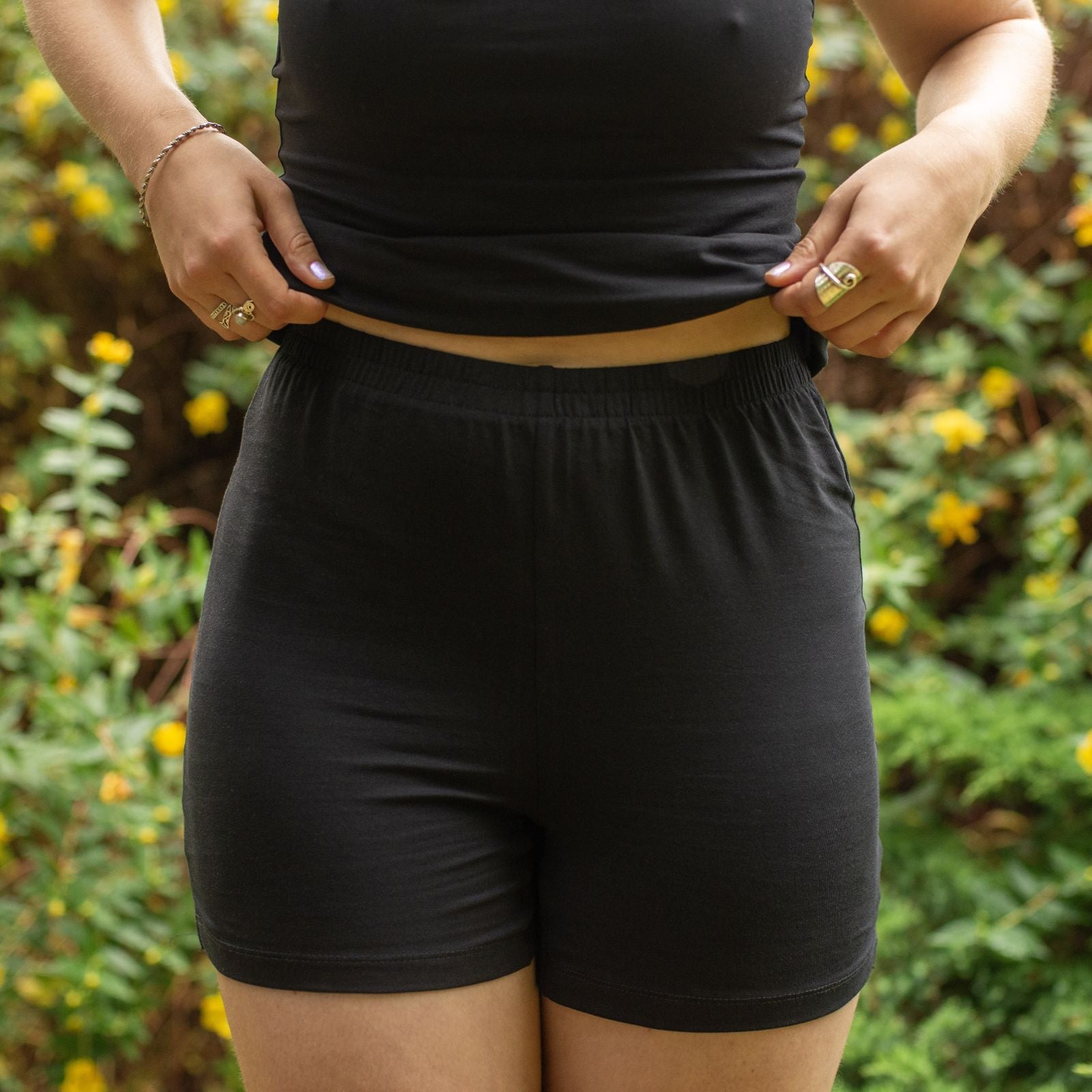 Product focused image of a model wearing the YOU Underwear Organic Cotton Black Undershorts. The model holds up her top with her hands, and stands in front of a bush with yellow flowers. The image is cropped from chest to upper thigh.