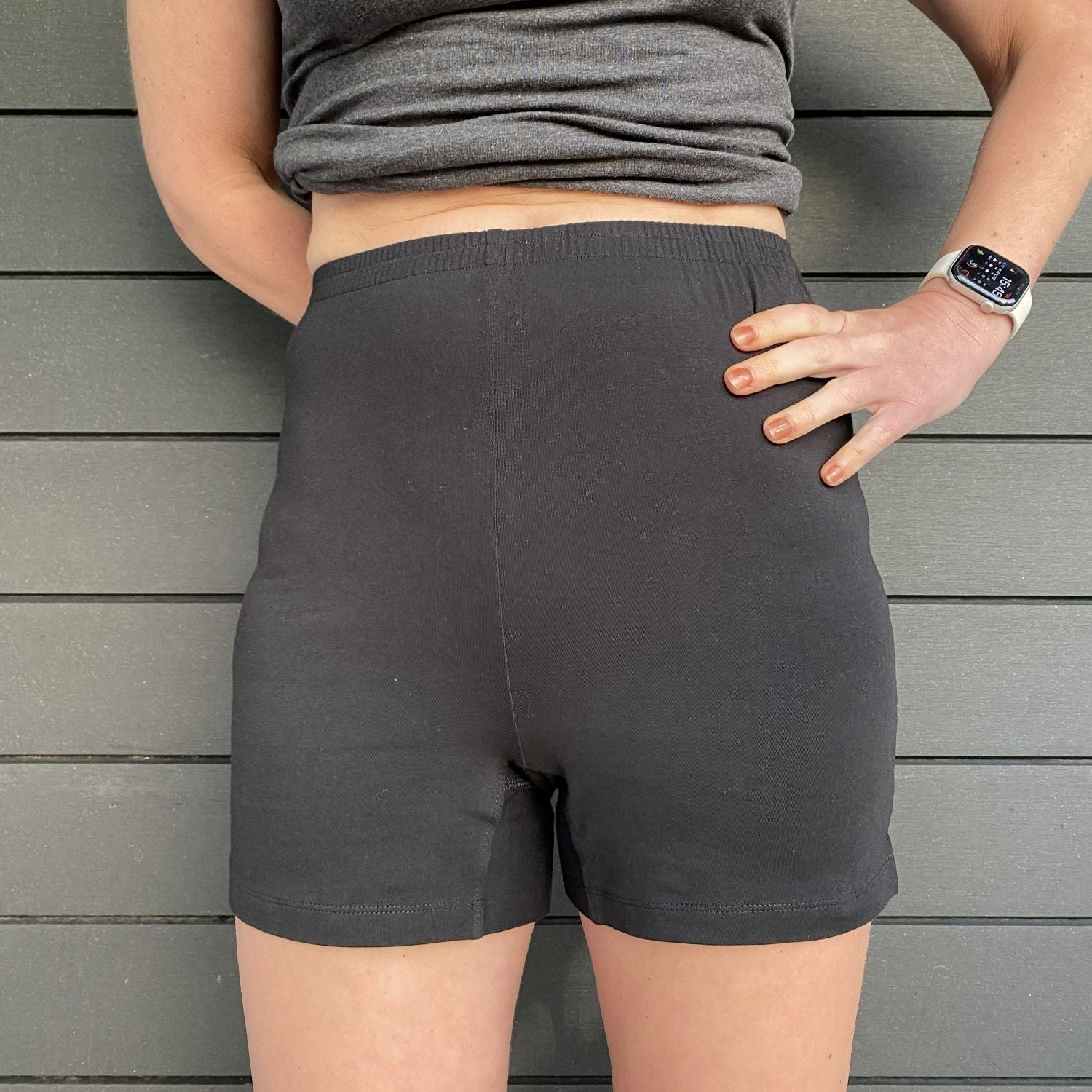 A close up photo of a model wearing the YOU Underwear undershorts in black. She is facing the camera, with her left hand on her hip. There is a grey paneled background and the photo is zoomed in so we can see from the model's mid-stomach to their mid-thigh.