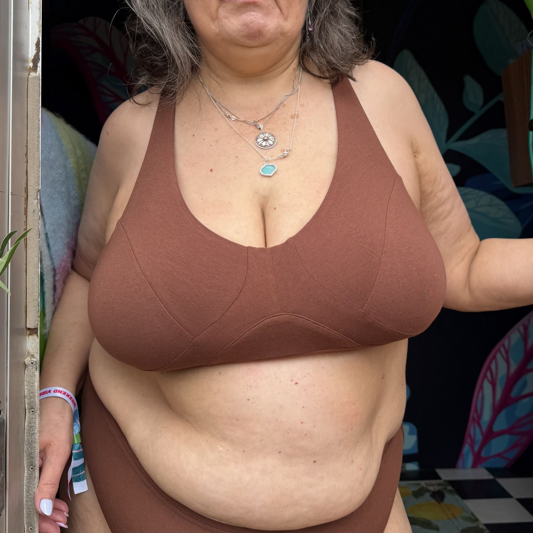 A photo of a model wearing the YOU Underwear non-wired bra and knickers in the colour chestnut. The photo is zoomed in so we can see from the model's shoulder's to their hips as they are facing the camera. The background is dark but there there is a wall painted with large leaves.