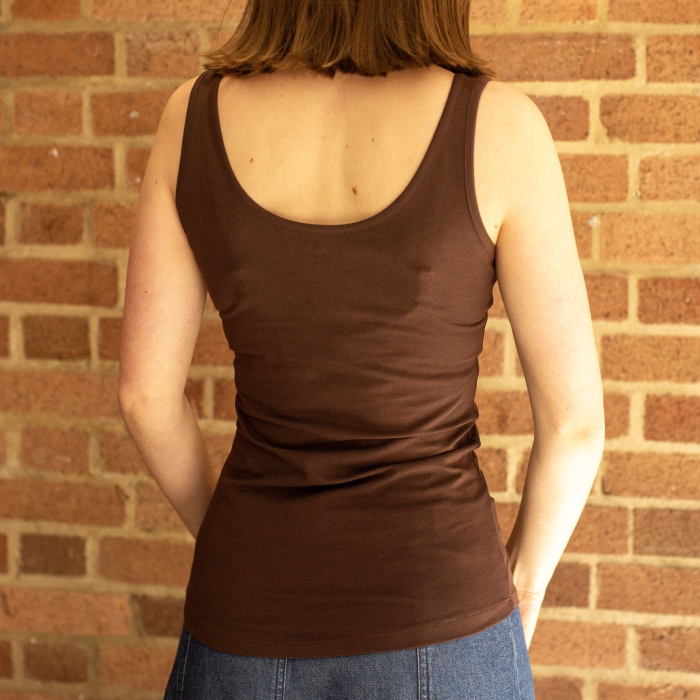 A product focused photo of a model wearing the YOU Underwear vest in cocoa. She is standing facing away from the camera, with her hands in front of her. The photo is zoomed in so we can see from her shoulders to her hips. There is a red brick wall behind her.