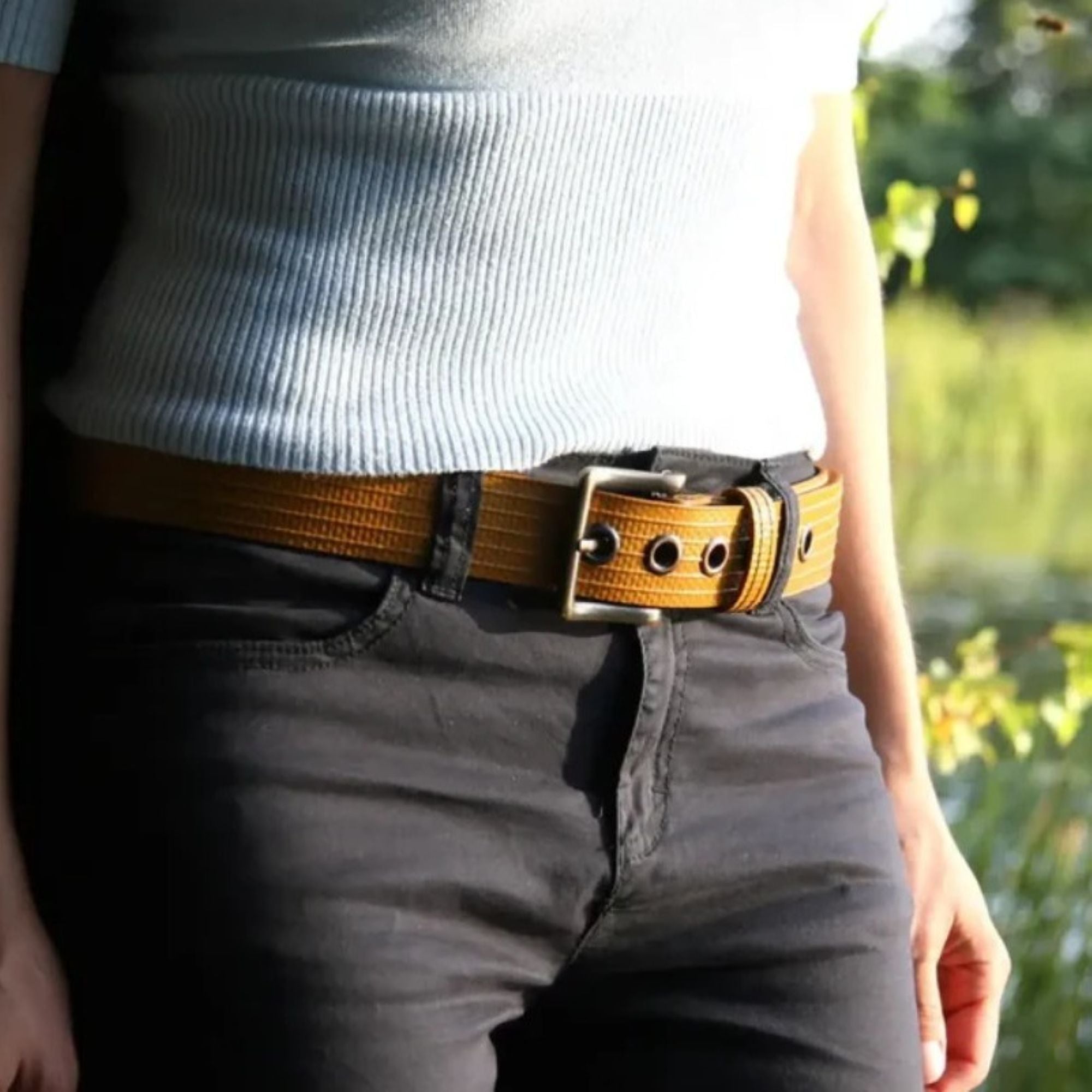A close up photo of a model wearing the Elvis & Kresse big yellow belt paired with a grey rib knit t-shirt and black jeans. The model is standing in front of a green garden.