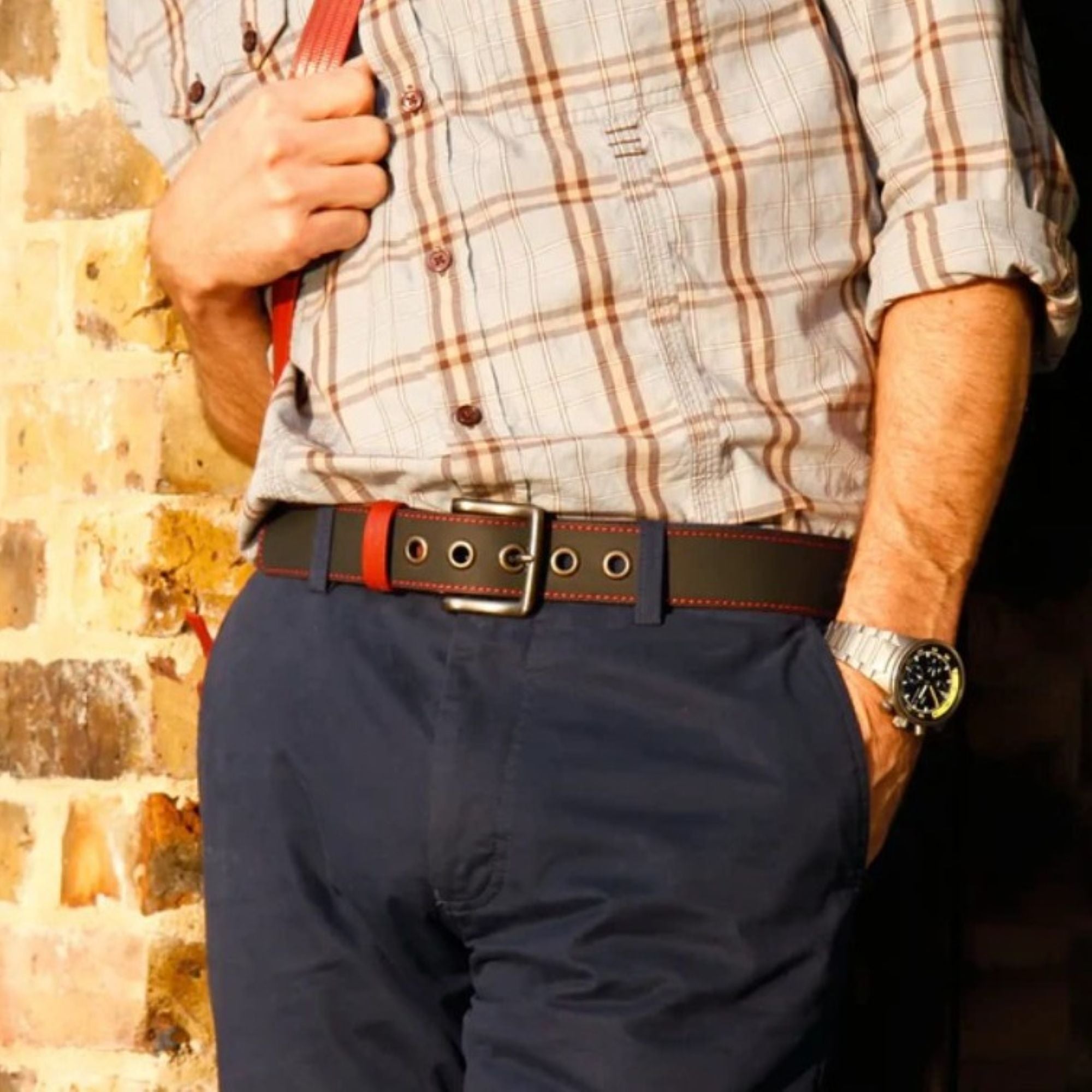 A close up of a model wearing the Elvis & Kresse black printing blanket belt paired with a red and grey checked shirt, dark blue jeans and a red shoulder bag.