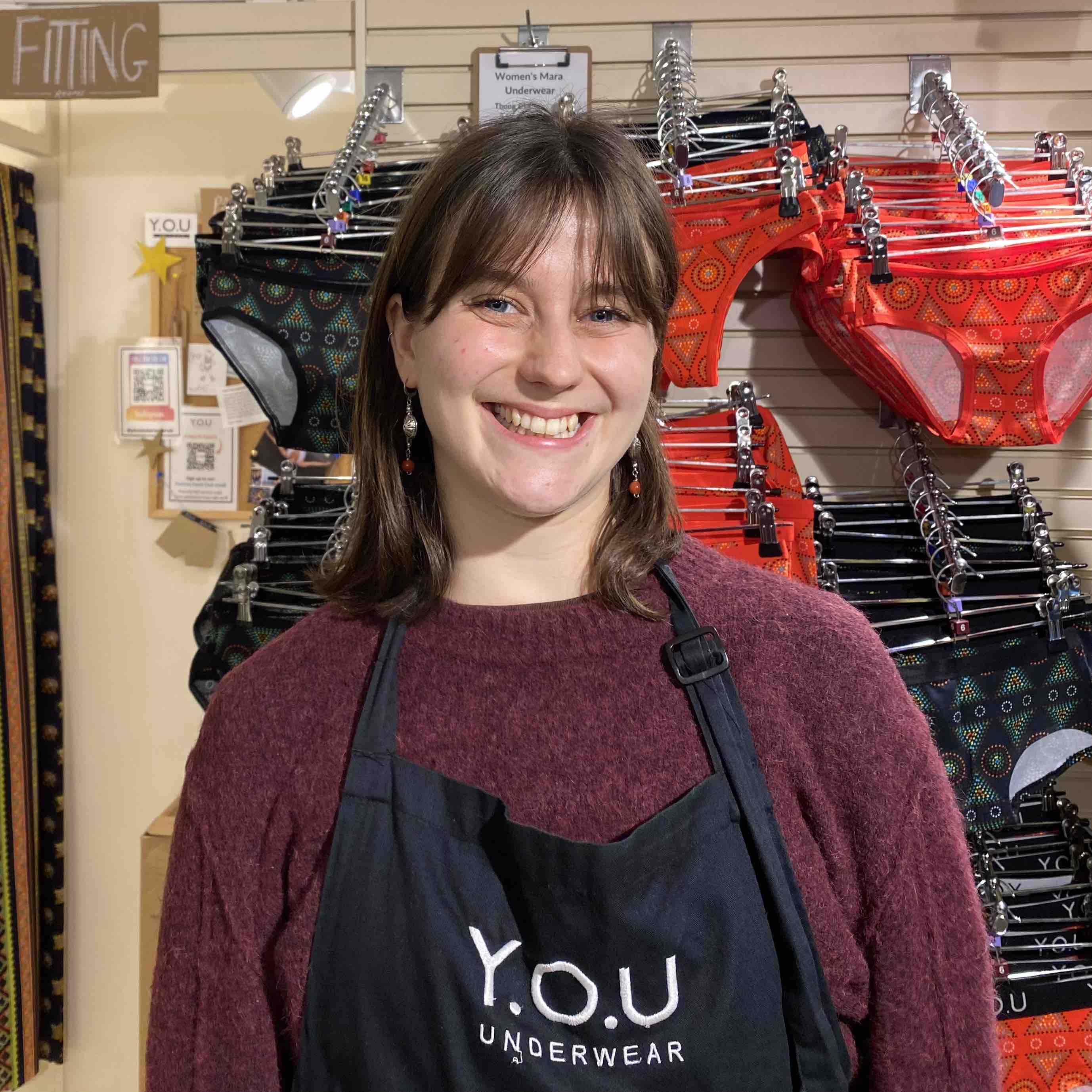 portrait photo of a woman wearing a red jumper and black apron, standing in front of a display of YOU mara underwear.
