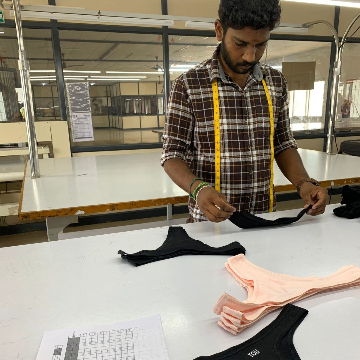 A photo of a man carrying out quality control checks to YOU Underwear black thongs. He is wearing a plaid shirt with a yellow tape measure around this neck. He standing behind a white table.
