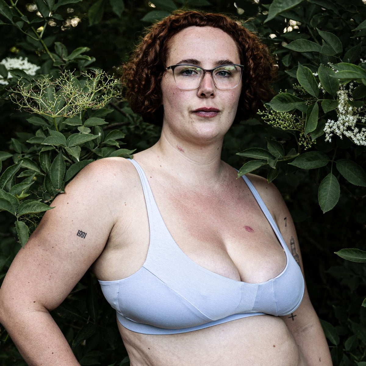 A photo of a model wearing the YOU Underwear non wired bra in light blue. She is standing facing the camera, with her right hand on her hip. She is surrounded by a dark green bush and the photo is zoomed in so we can see from her head to her mid-stomach.