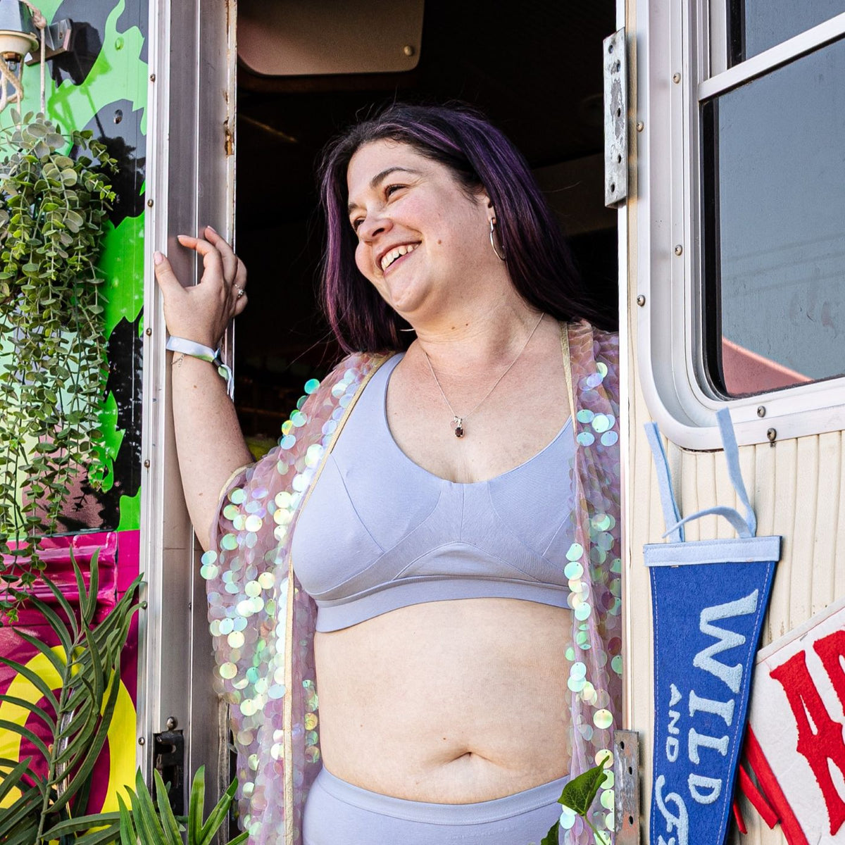 A photo of a model wearing the YOU Underwear non-wired bra and boyshorts in light blue. She is standing in the doorway of a bus with her hands on the door frame and looking out smiling. She is wearing a light pink, sparkly cover up. There are hanging plants to her left and bright blue and red empowering flags on her right.