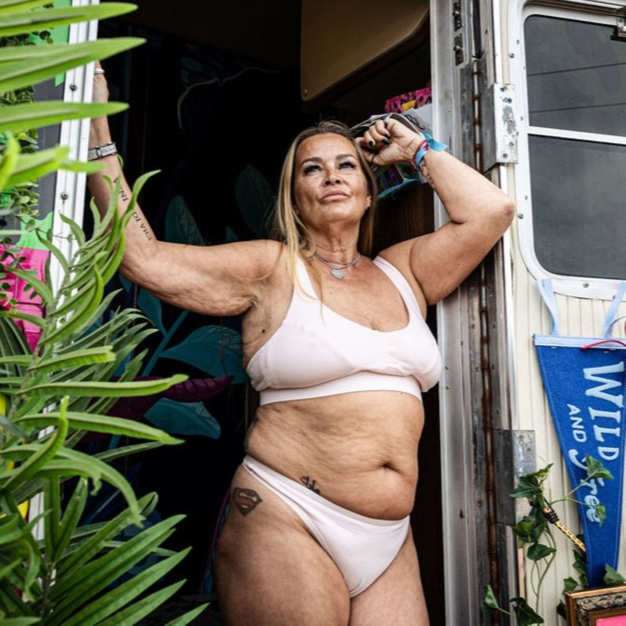 A photo of a model wearing the YOU Underwear light pink non-wired bra and knickers. She is standing in the doorway of a camper-van, leaning against the frame. She is looking out into the distance with a slight pout on her face. There is green foliage in the left foreground of the photo.