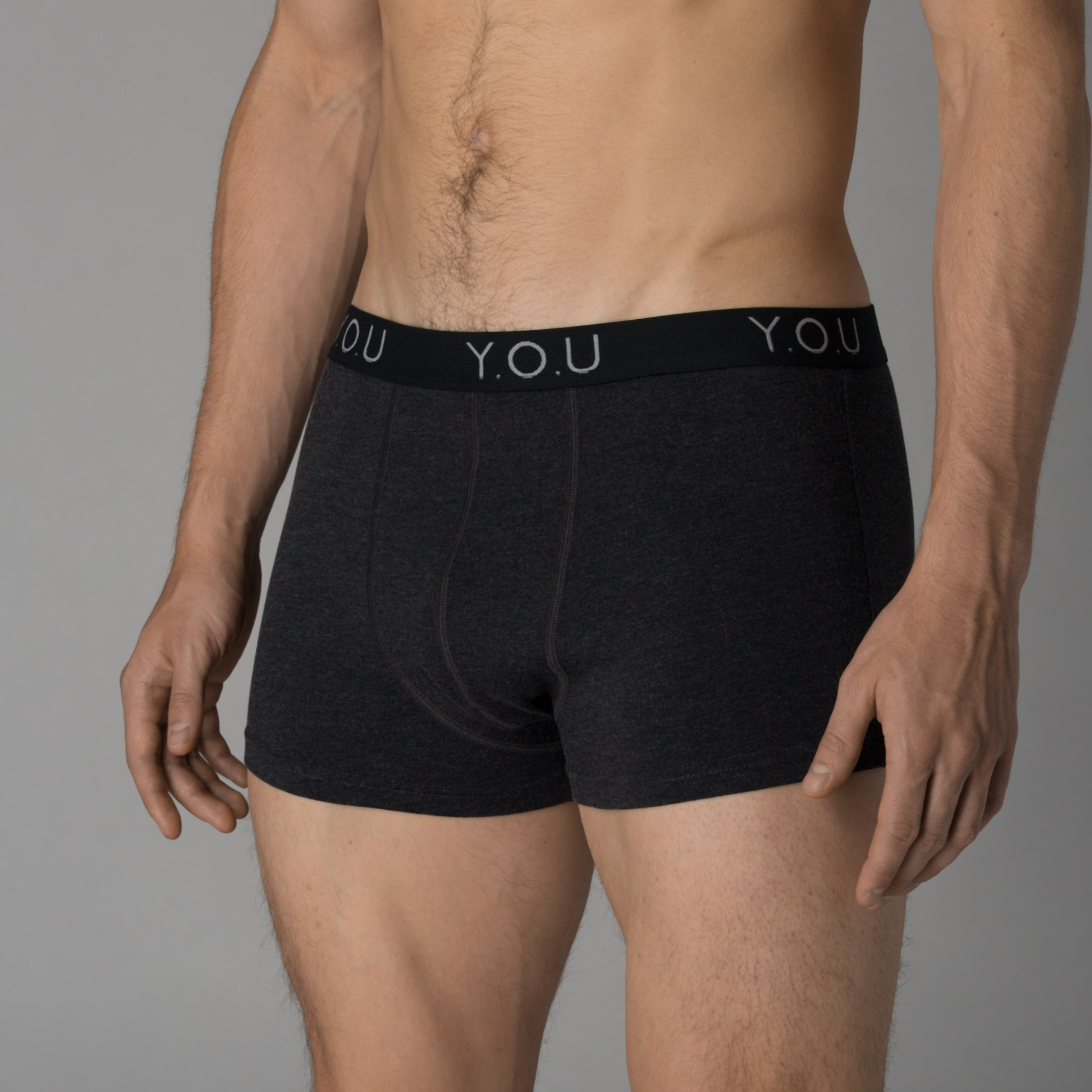 A man is standing against a plain background, wearing dark grey hipster briefs. The waistband displays the brand name "Y.O.U". The image focuses on his torso and upper legs, with his hands held loosely at his sides.
