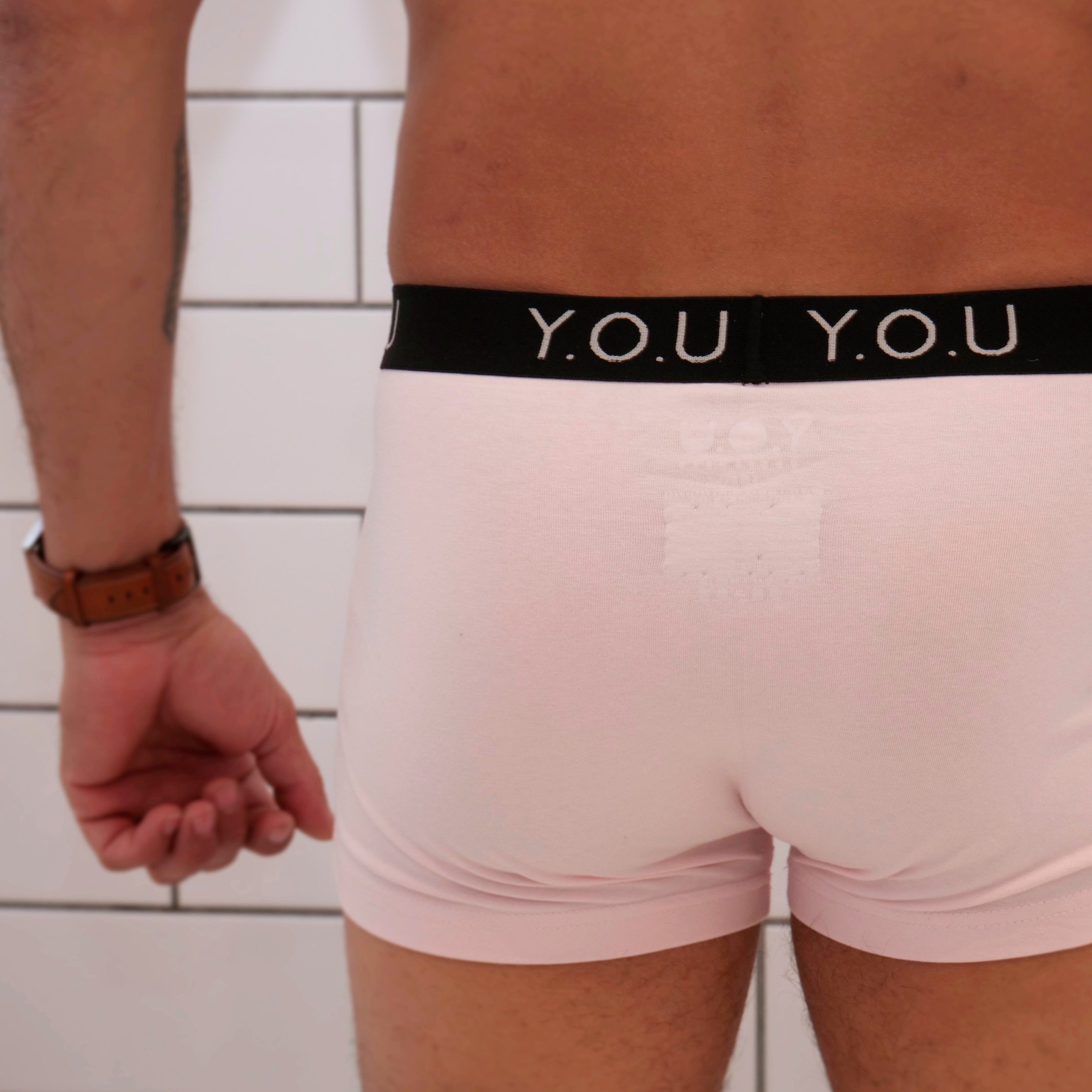A close-up of a person from behind wearing light pink men's hipster trunks with a black waistband bearing the letters "Y.O.U."