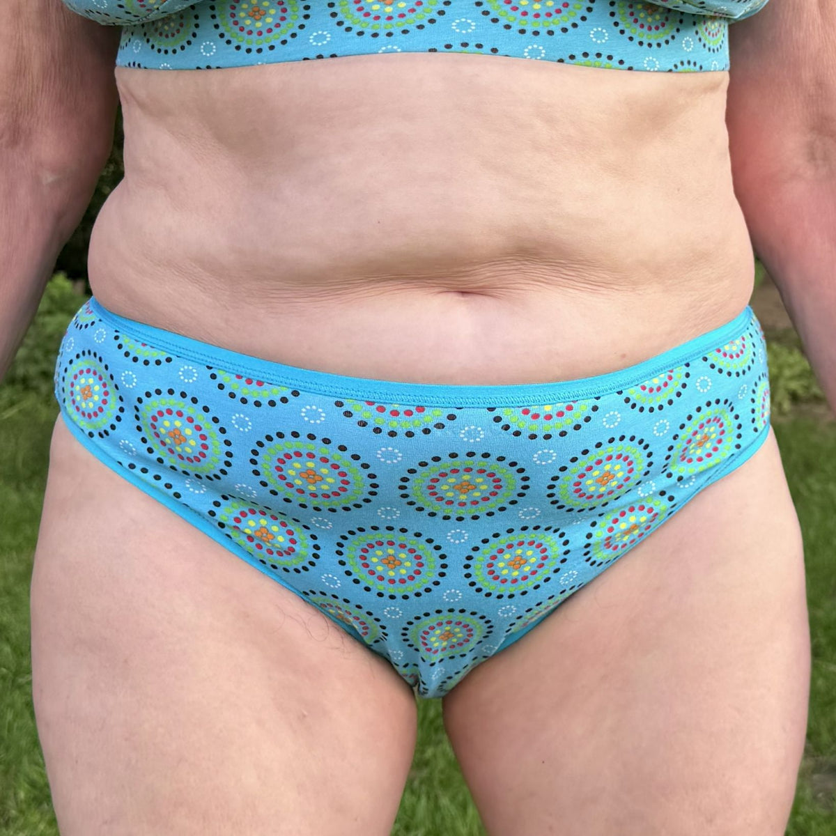 A close up photo of a model wearing the YOU Underwear mid rise bikini knickers in the blue mara print. They are standing facing the camera with their hands at their sides. We can see from the model's stomach to their upper-thigh. There is a green field in the background.