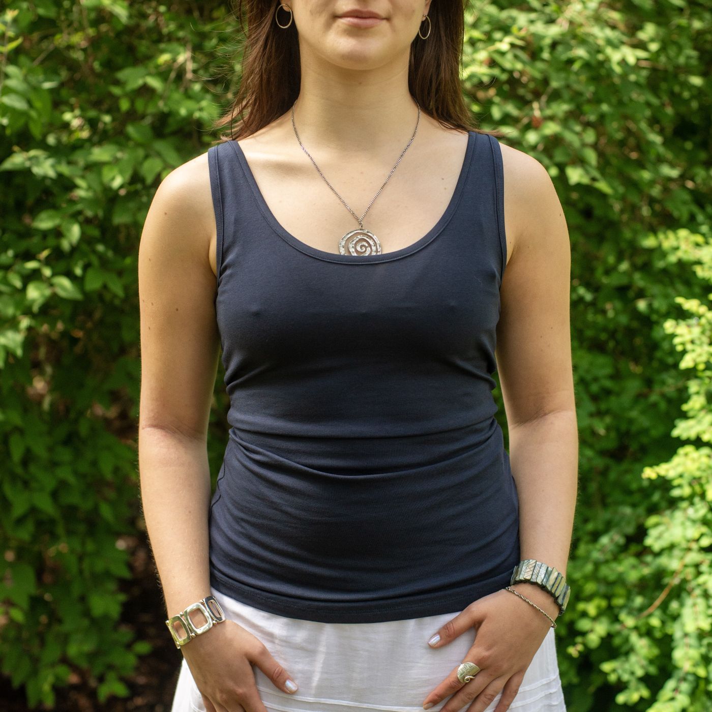 A close up of a model wearing the YOU Underwear navy vest. She is facing the camera, with her hands at her sides. She is standing in front of green bush.
