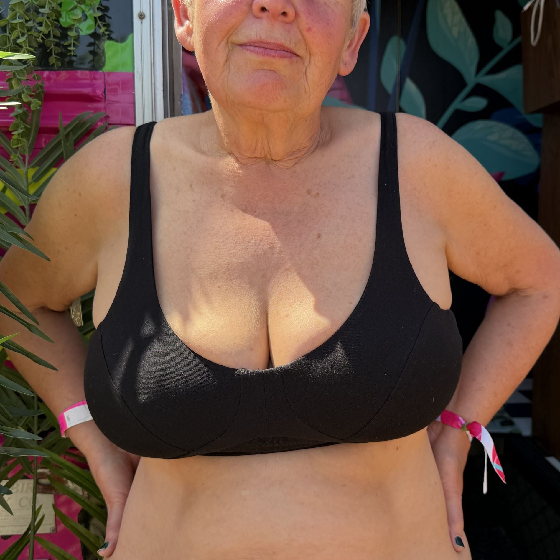 A photo of a model wearing the YOU Underwear non-wired bra in black. The photo is zoomed in so we can see from the model's nose to their mid-stomach. They are standing with their hands on their hips, with plants behind them on their left.
