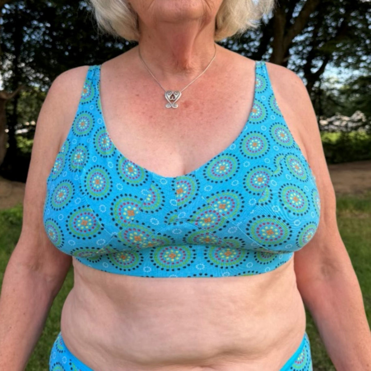 A photo of a model wearing the YOU Underwear non wired bra in the bright blue mara print. They are standing facing the camera with their hands at their sides. The photo is zoomed in so we can only see from the model's shoulders to their waist. There is a green and woody field in the background.