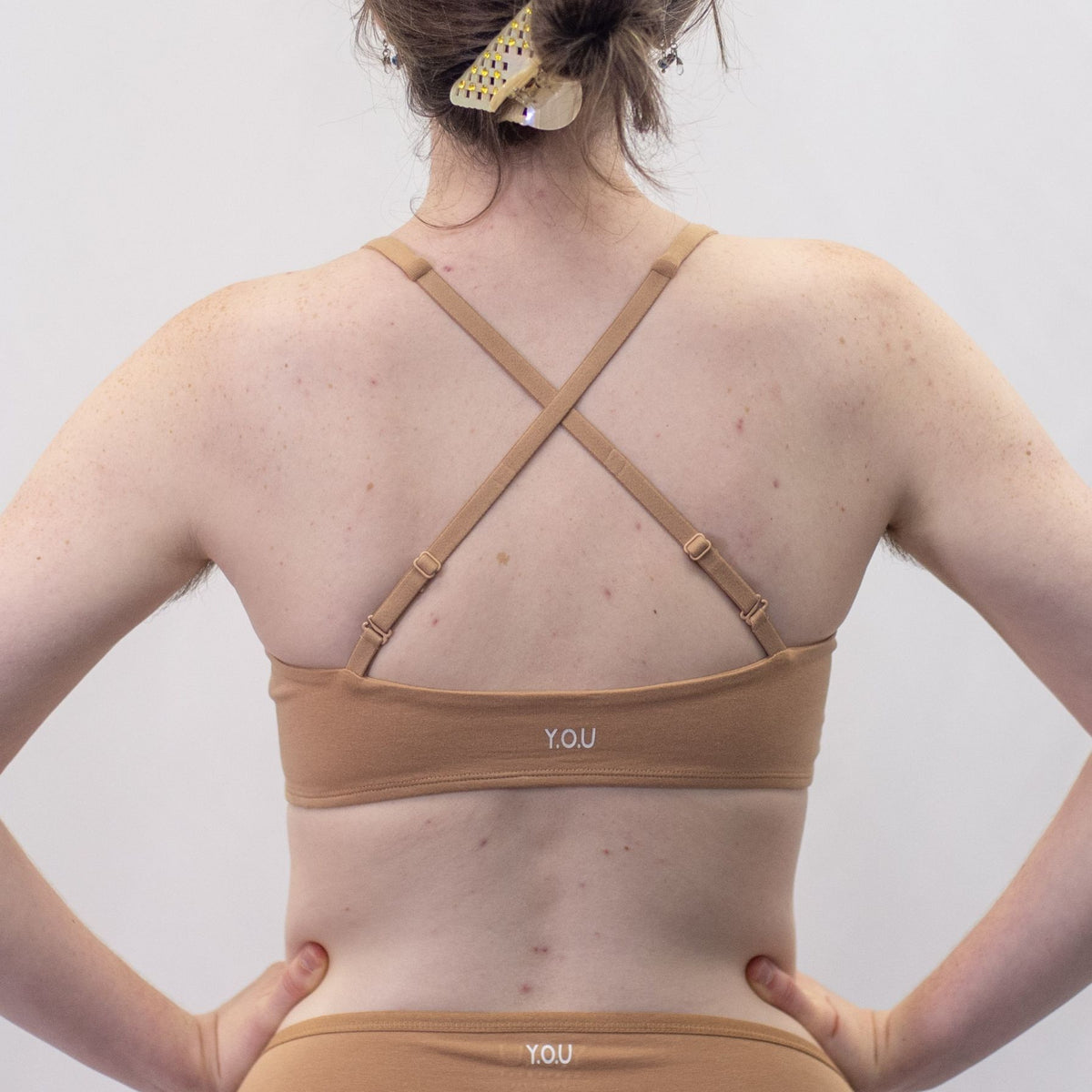 A photo of a model wearing the YOU Underwear padded bralette in almond. She is standing facing away from the camera with her hands on her hips. The photo is cropped so we can see from the model's shoulders to their waist. There is a white background.