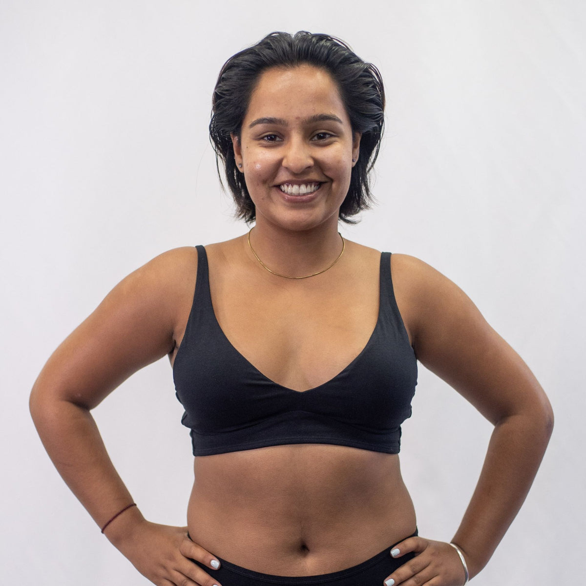 A photo of a model wearing the YOU Underwear padded bralette in black. They are standing facing the camera with their hands on their hips. The photo is cropped so we can only see to the model's waist. There is a white background.
