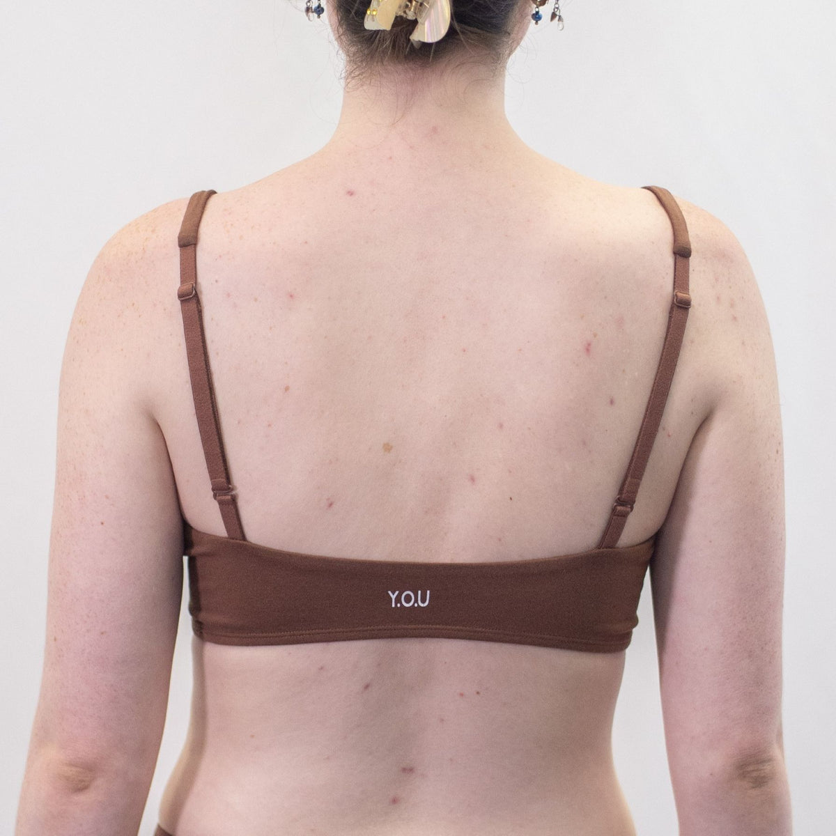 A photo of a model wearing the YOU Underwear padded bralette in chestnut. She is standing facing away from the camera with both hands at her side. The photo is cropped so we can see from the model's chin to their mid-stomach. There is a white background.