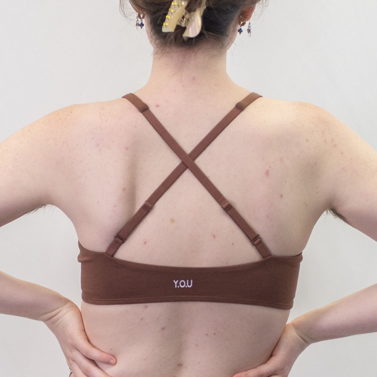 A photo of a model wearing the YOU Underwear padded bralette in chestnut. She is standing facing away from the camera with both hands on her hips. The photo is cropped so we can see from the model's ears to their mid-back. There is a white background.