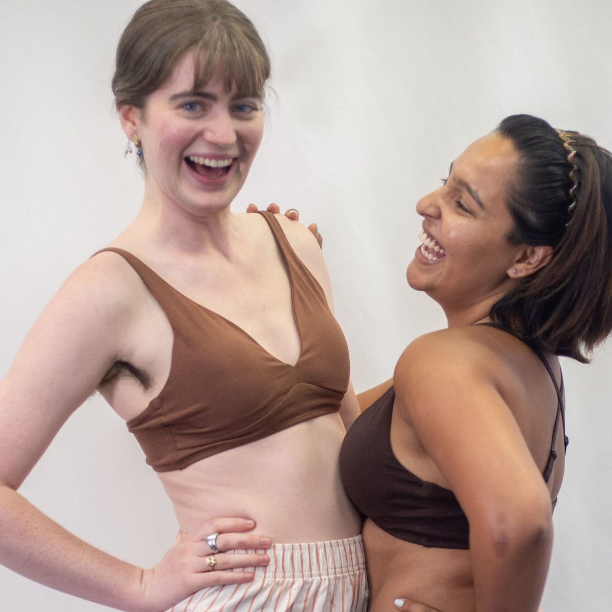 A photo of two models one wearing the YOU Underwear padded bralette in chestnut and one wearing it in cocoa. They are embraced together and laughing, one looking at the camera and another looking at the other. There is a white background.