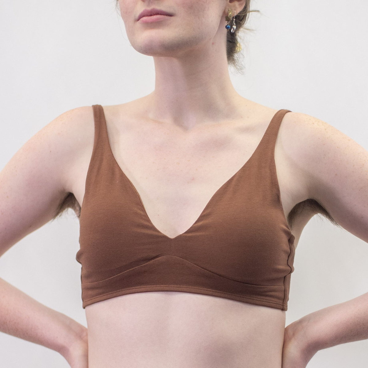 A photo of a model wearing the YOU Underwear padded bralette in chestnut. She is standing facing the camera with both hands on her hips. The photo is cropped so we can see from the model's chin to their mid-stomach. There is a white background.