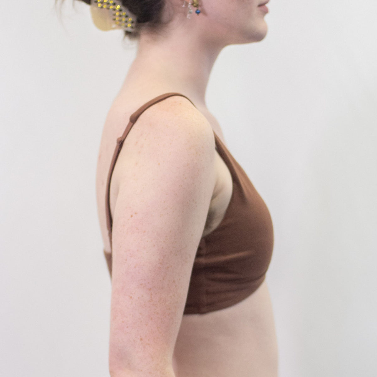 A photo of a model wearing the YOU Underwear padded bralette in chestnut. She is standing side on to the camera with both hands on her hips. The photo is cropped so we can see from the model's chin to their mid-stomach. There is a white background.