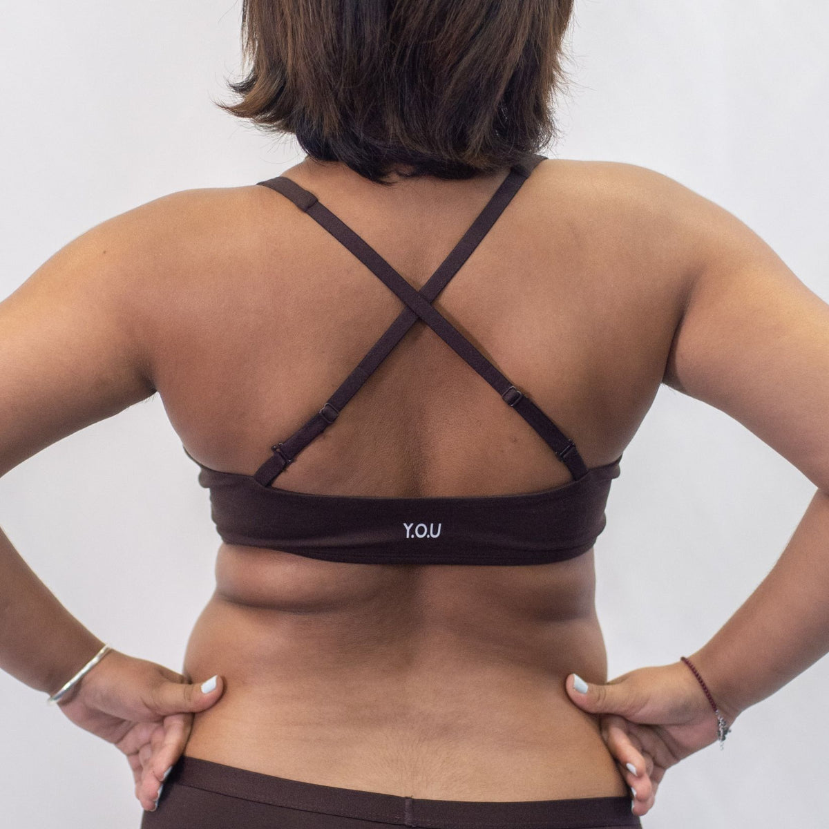 A photo of a model wearing the YOU Underwear padded bralette in cocoa. She is standing facing away from the camera with both hands on her hips, smiling. The photo is cropped so we can see from the model's shoulders to their hips. The straps of the bralette are crossed to show another style of wear. There is a white background.