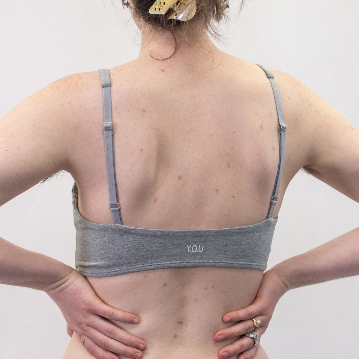 A photo of a model wearing the YOU Underwear padded bralette in grey. They are standing facing away from the camera with their hands on their hips. The photo is zoomed in so we can see from the model's shoulders to their mid-stomach. There is a white background.