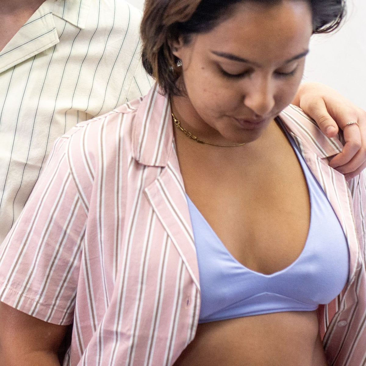 A photo of a model wearing the YOU Underwear padded bralette in light blue paired with an open stripey shirt. The photo is cropped so we can see the shoulder of another model, wearing a cream and blue striped shirt who has one arm around the model wearing the blue bralette.