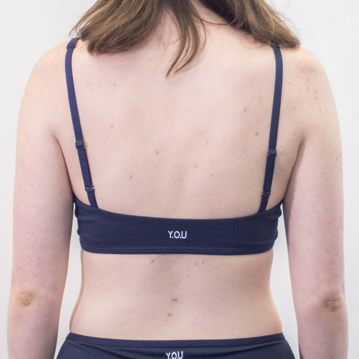 A photo of a model wearing the YOU Underwear padded bralette in navy blue. She is standing facing away from the camera and the photo is cropped so we can see from her shoulders to her waist. There is a white background.