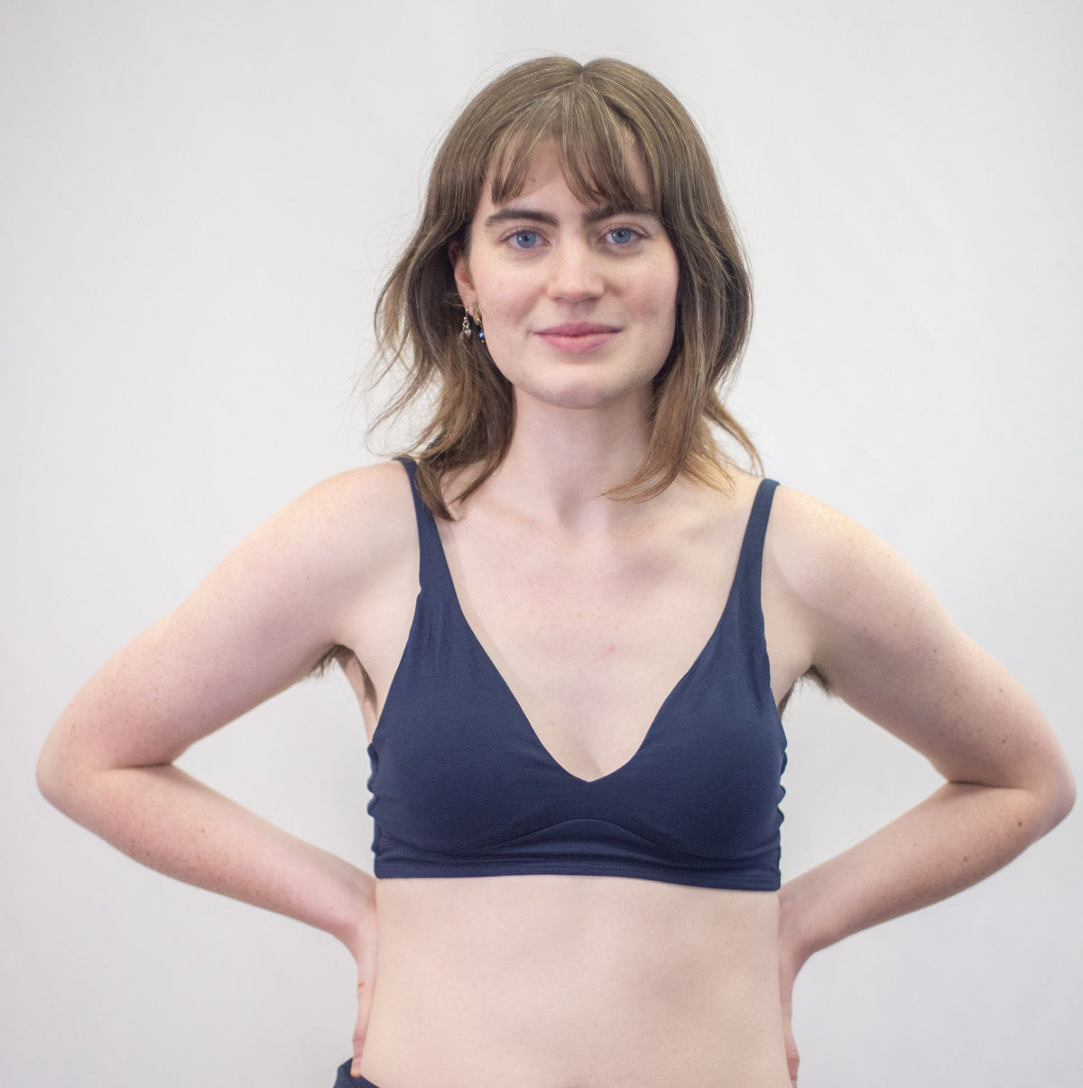 A photo of a woman with shoulder length brown hair wearing the YOU Underwear padded bralette in navy blue. They are standing facing the camera with their hands on their hips. The photo is zoomed in so we can see the model down to just above their belly button. There is a white background.
