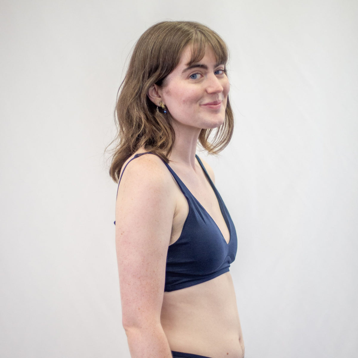 A photo of a model wearing the YOU Underwear padded bralette in navy blue. She is standing side on to the camera with her hands at her sides, looking at the lens. There is a white background and the photo is cropped so we can see to the model's waist.