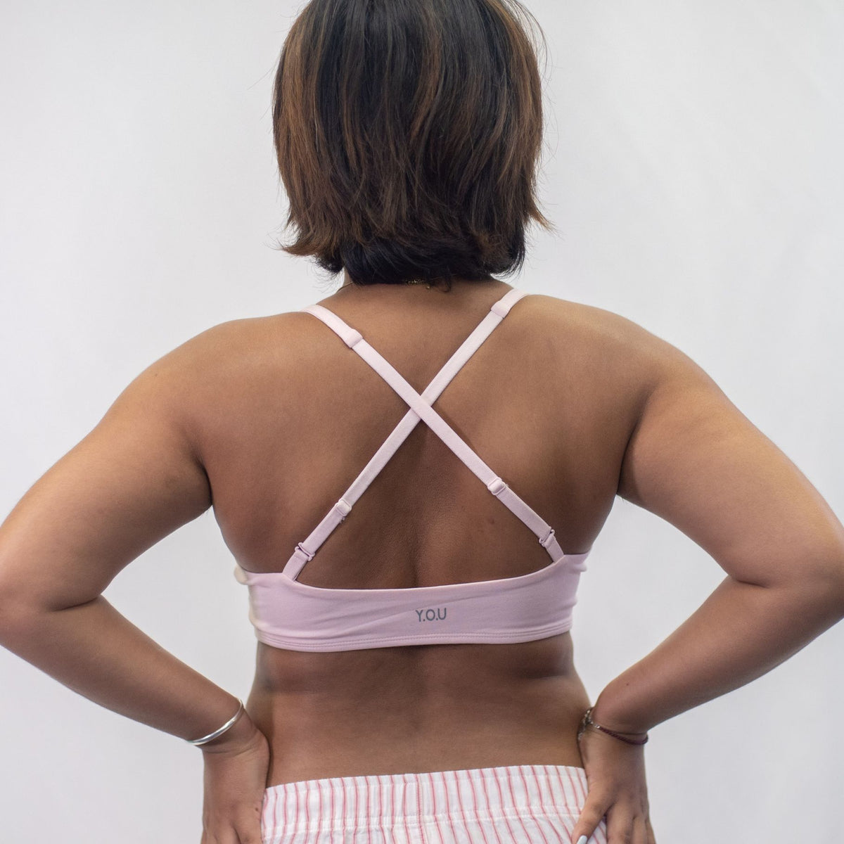 A photo of a model wearing the YOU Underwear padded bralette in pink. She is standing facing away from the camera with both hands on her hips. The straps of the bralette are crossed to show a different style of wear. The photo is cropped so we can see to the model's hips. There is a white background.