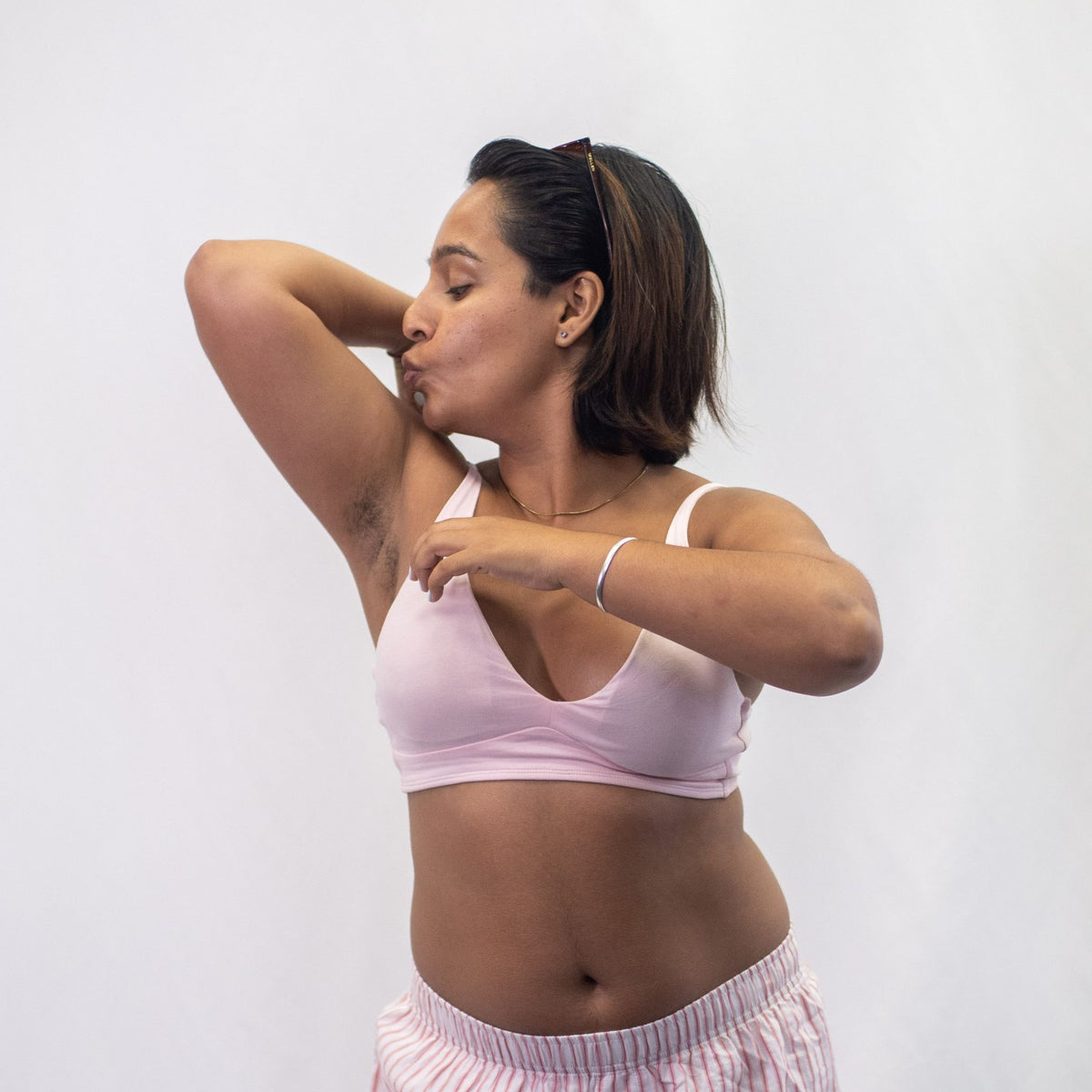 A photo of a model wearing the YOU Underwear padded bralette in pink. She is standing facing the camera and kissing her right flexed bicep. The photo is cropped so we can see to the model's hips. There is a white background.