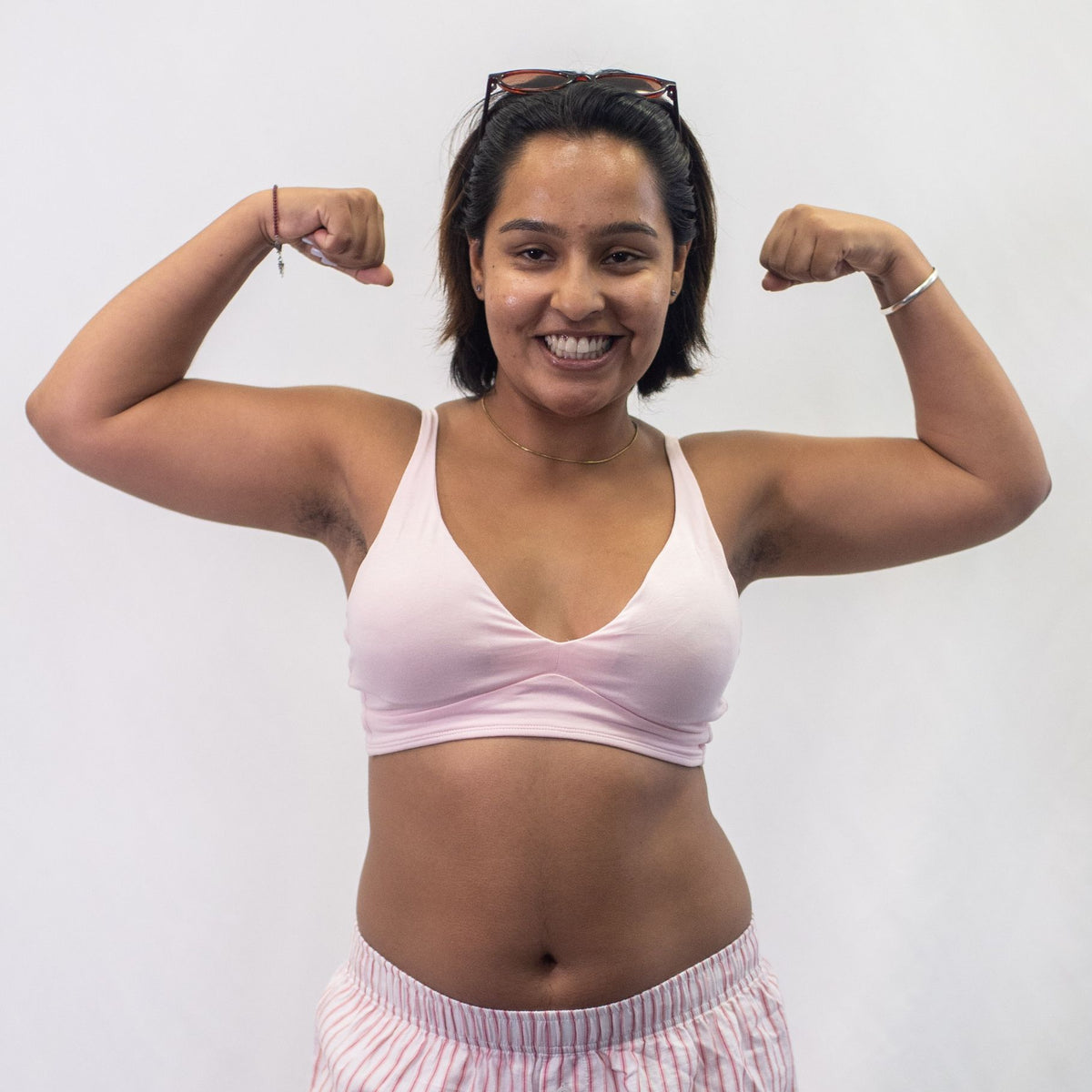 A photo of a smiling woman posing with her arms in the air wearing the YOU Underwear padded bralette in light pink. There is a white background.