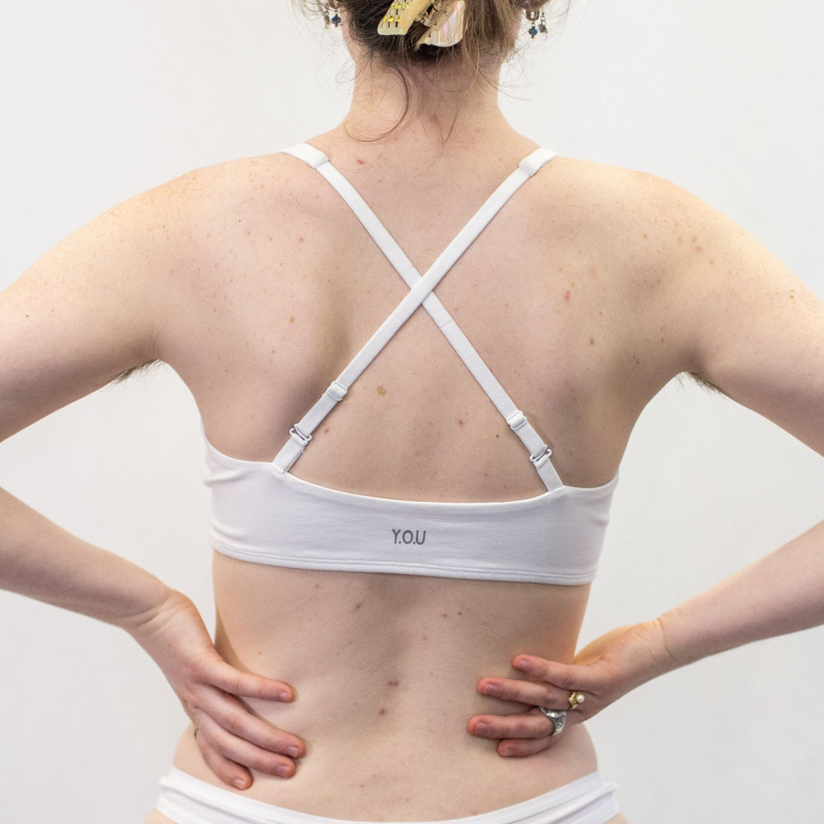 A photo of a model wearing the YOU Underwear bralette in white. They are standing facing away from the camera with their hands on their hips. The photo is cropped so we can see from the model's shoulders to their waist. The bralette has the straps crossed to show another style of wear. The background is white.