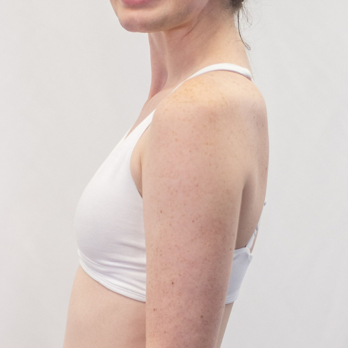 A photo of a model wearing the YOU Underwear bralette in white. They are standing side on to the camera with their hands at their sides. The photo is cropped so we can see from the model's shoulders to their mid-stomach. The background is white.