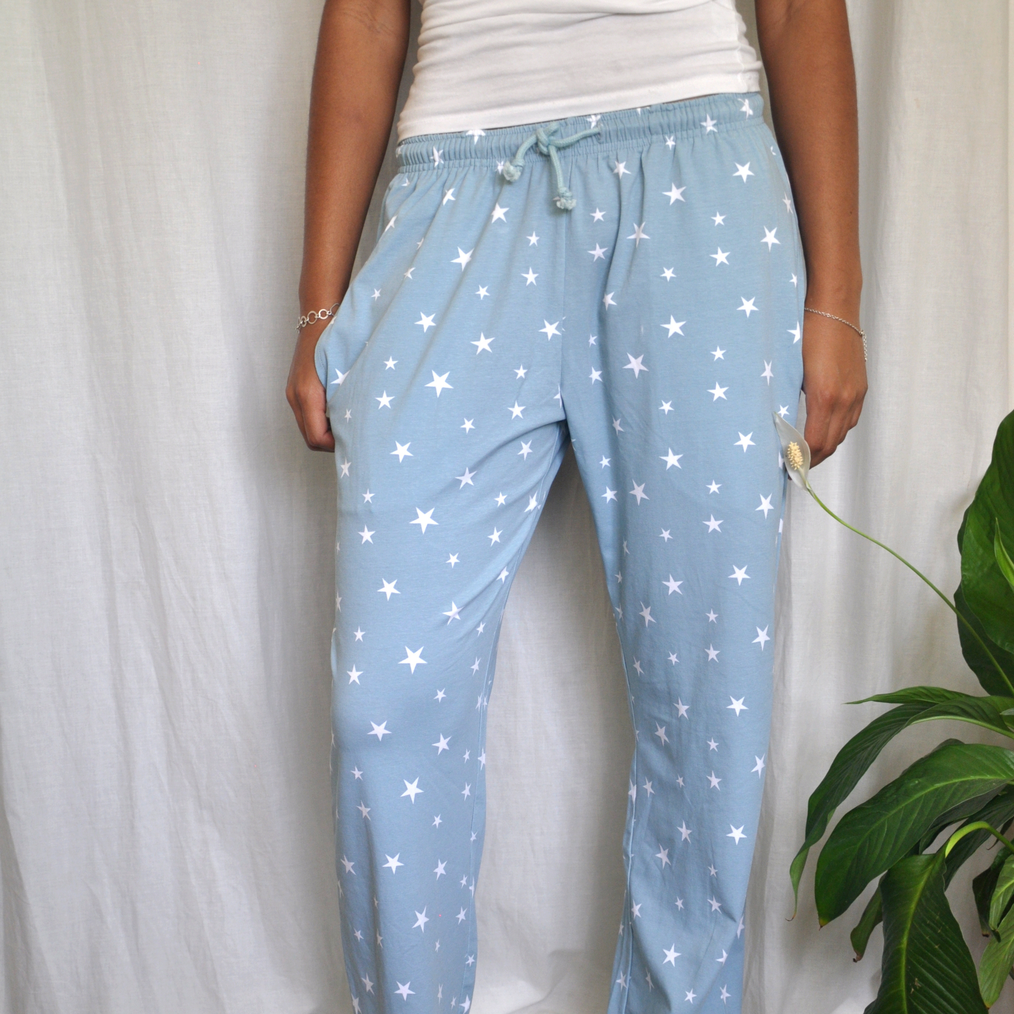 A person wearing pyjama bottoms in a blue with white stars pattern stands against a white curtain background. Their hands are casually placed in the pants' pockets. A green plant is partially visible to the right of the person.
