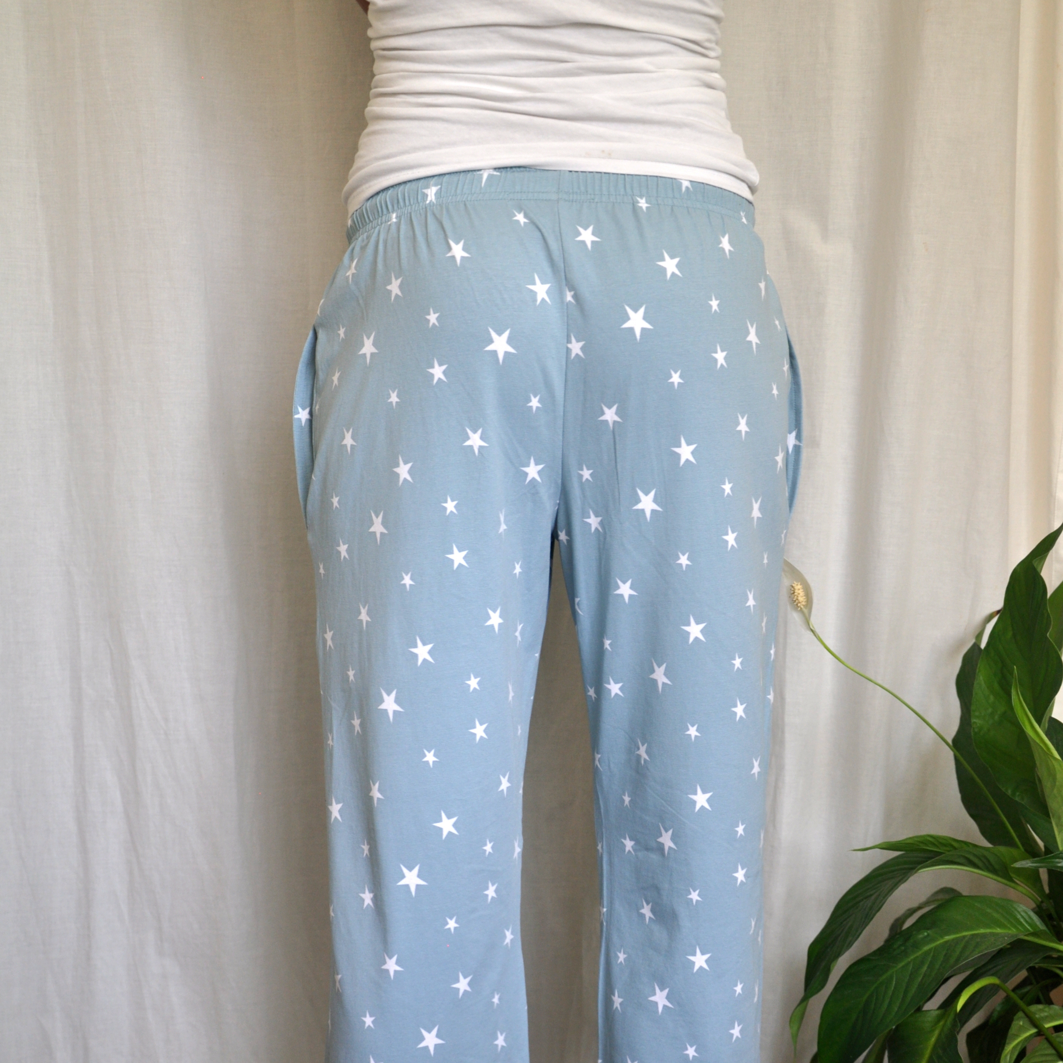 Person wearing light blue pajama bottoms with white star patterns, a white top, and standing with their back toward the camera. A green plant is visible on the right side in the bottom corner.