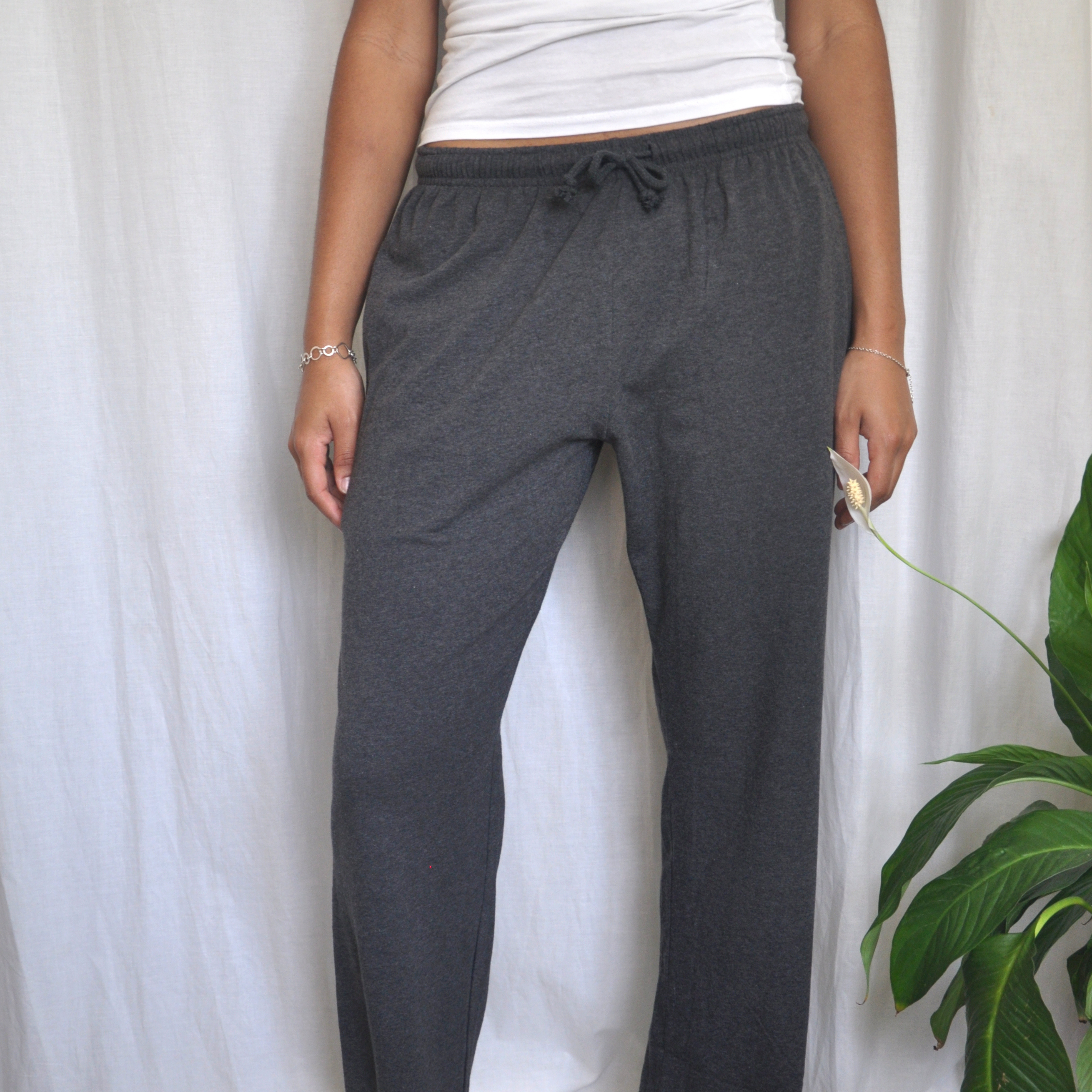 Person standing in front of a white curtain, wearing a white sleeveless top and dark grey drawstring pyjama pants. Their left hand, adorned with bracelets, rests on their side. On their right side, a green plant with long leaves is visible.